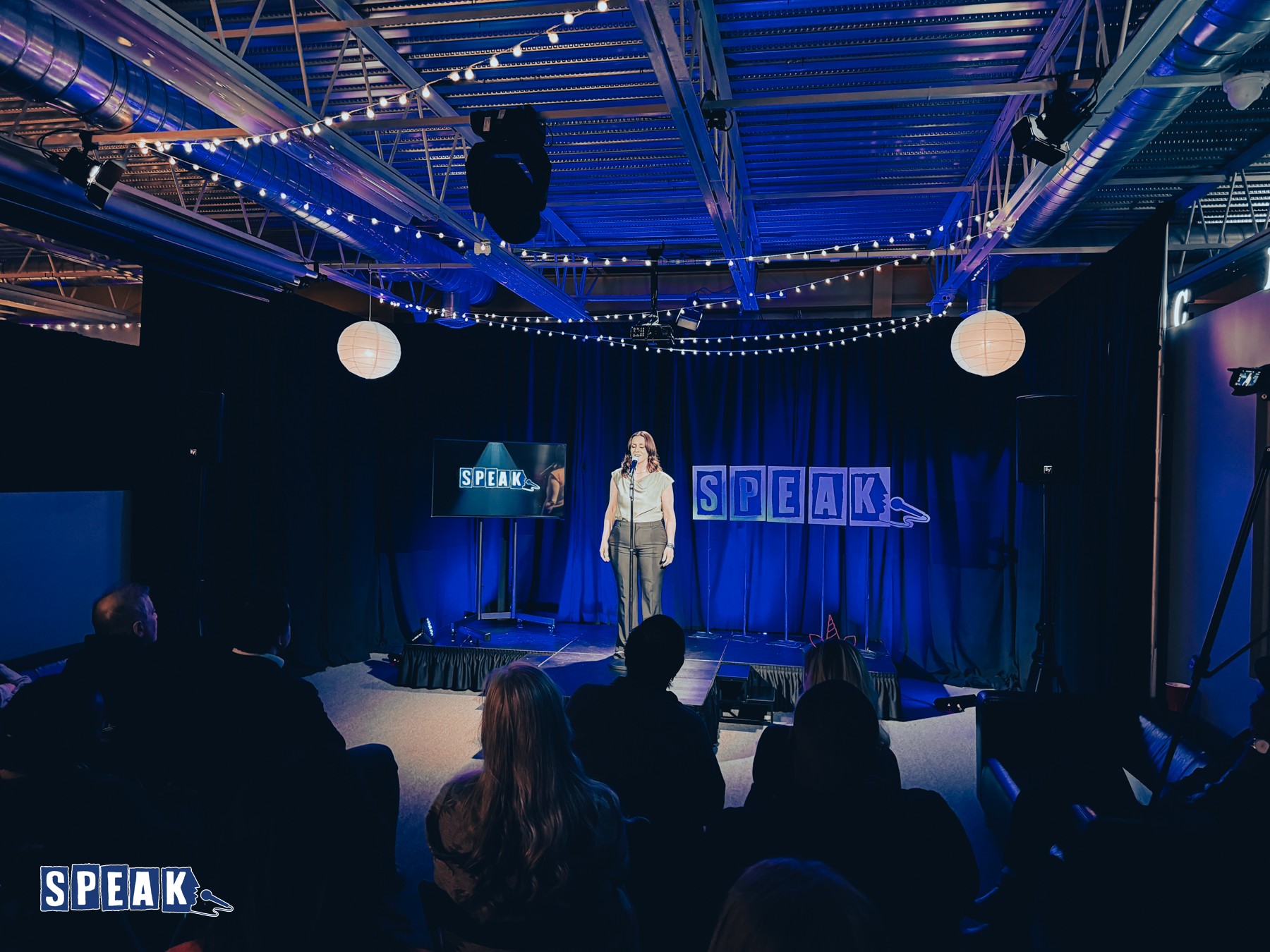 Person standing on stage with a microphone, audience seated watching, stage decorated with blue curtains and signs reading 'SPEAK'.