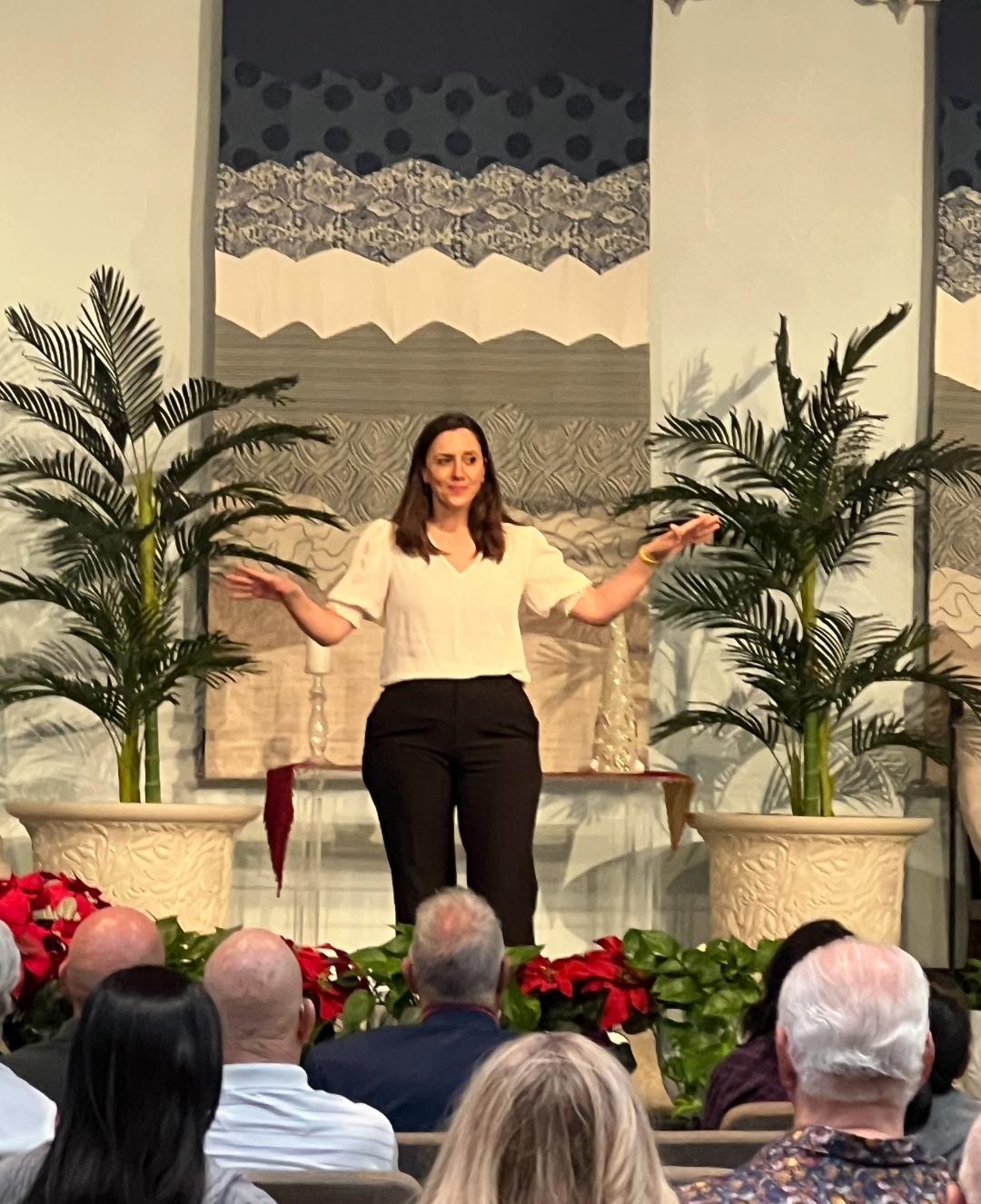 Woman standing on stage with arms outstretched, two large potted plants on either side, audience watching, decorated background.