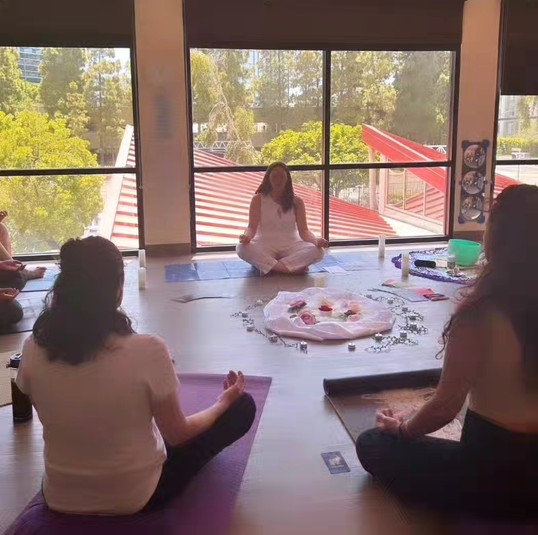 Three women practicing meditation in a bright room with large windows, sitting on mats around a tray of items.