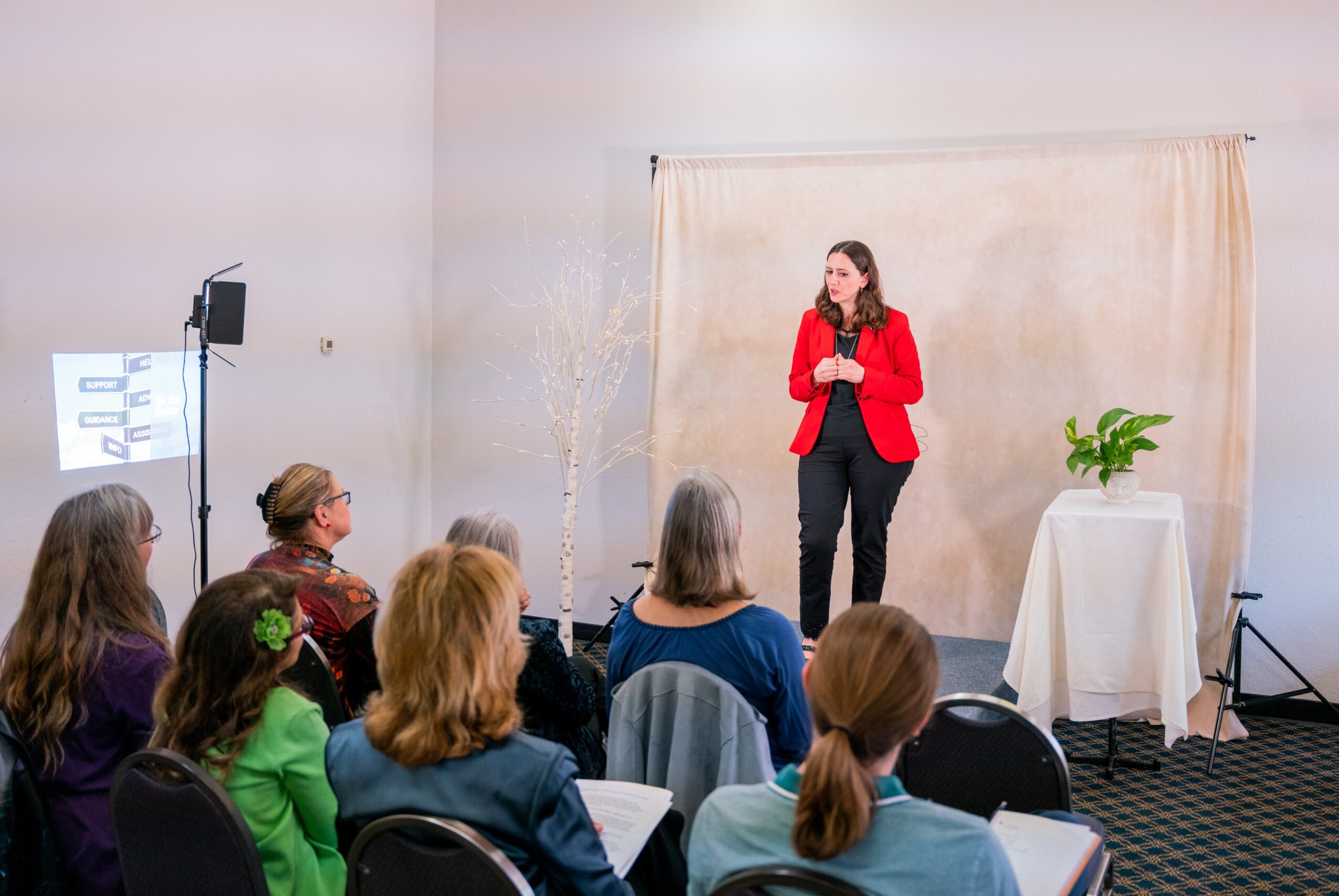Woman in red blazer speaking to an audience in a room with a beige backdrop and a potted plant on a table.