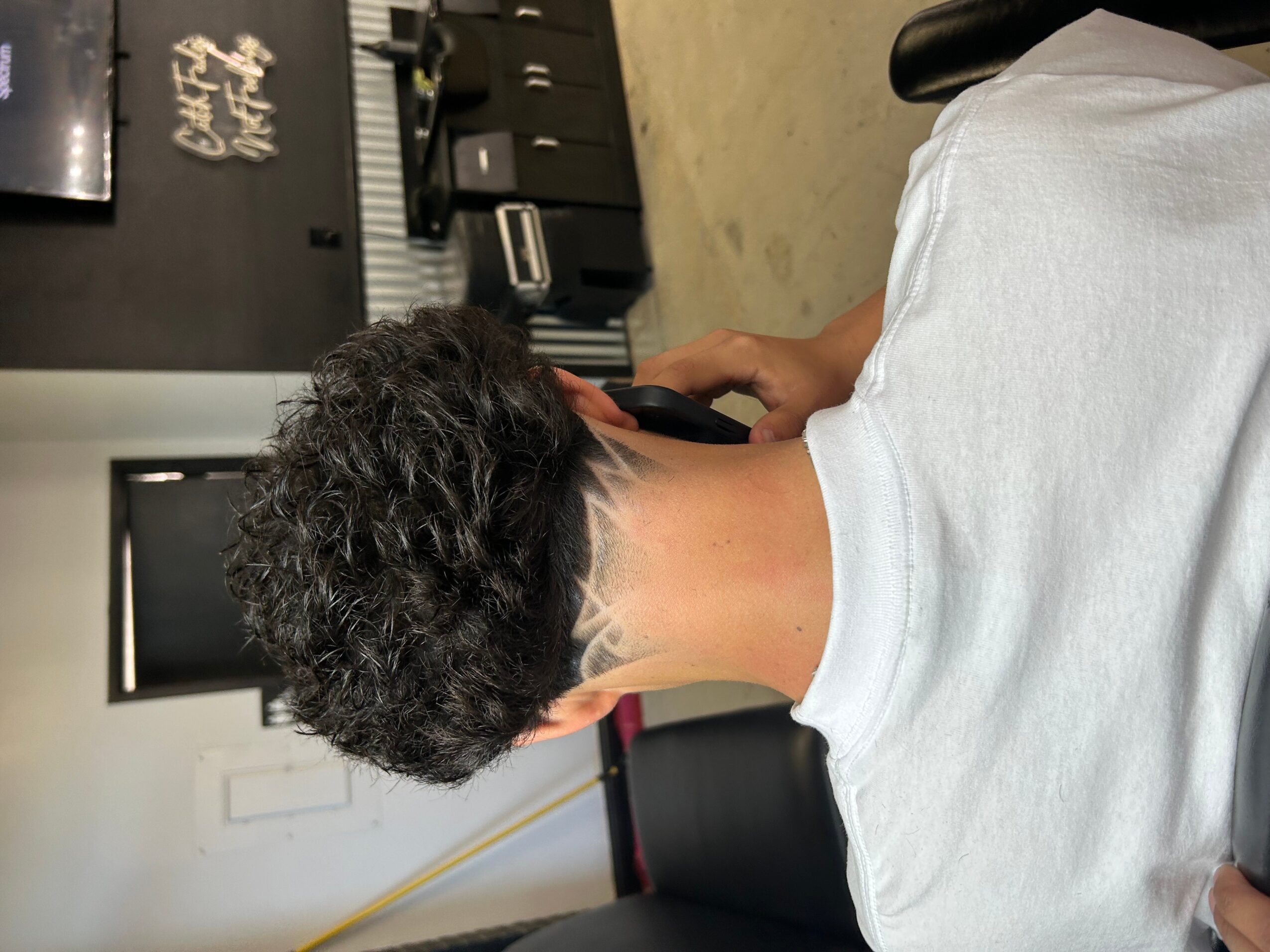 Person with short, curly hair sitting in a salon chair, facing away, with a white cape over their shoulders.