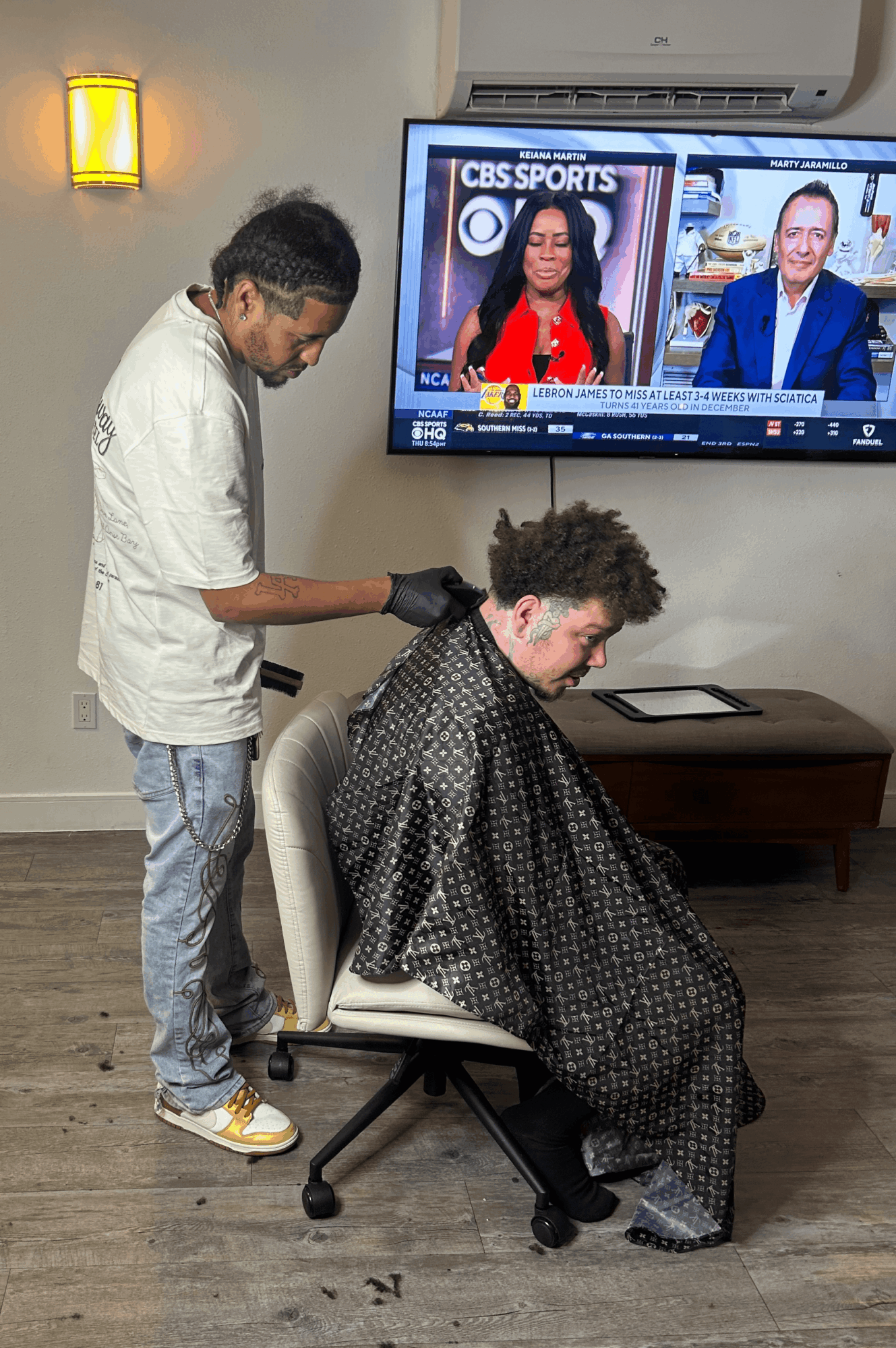 Person getting a haircut in a salon, seated in a chair with a cape, while a stylist works on their hair. TV screen shows sports news.