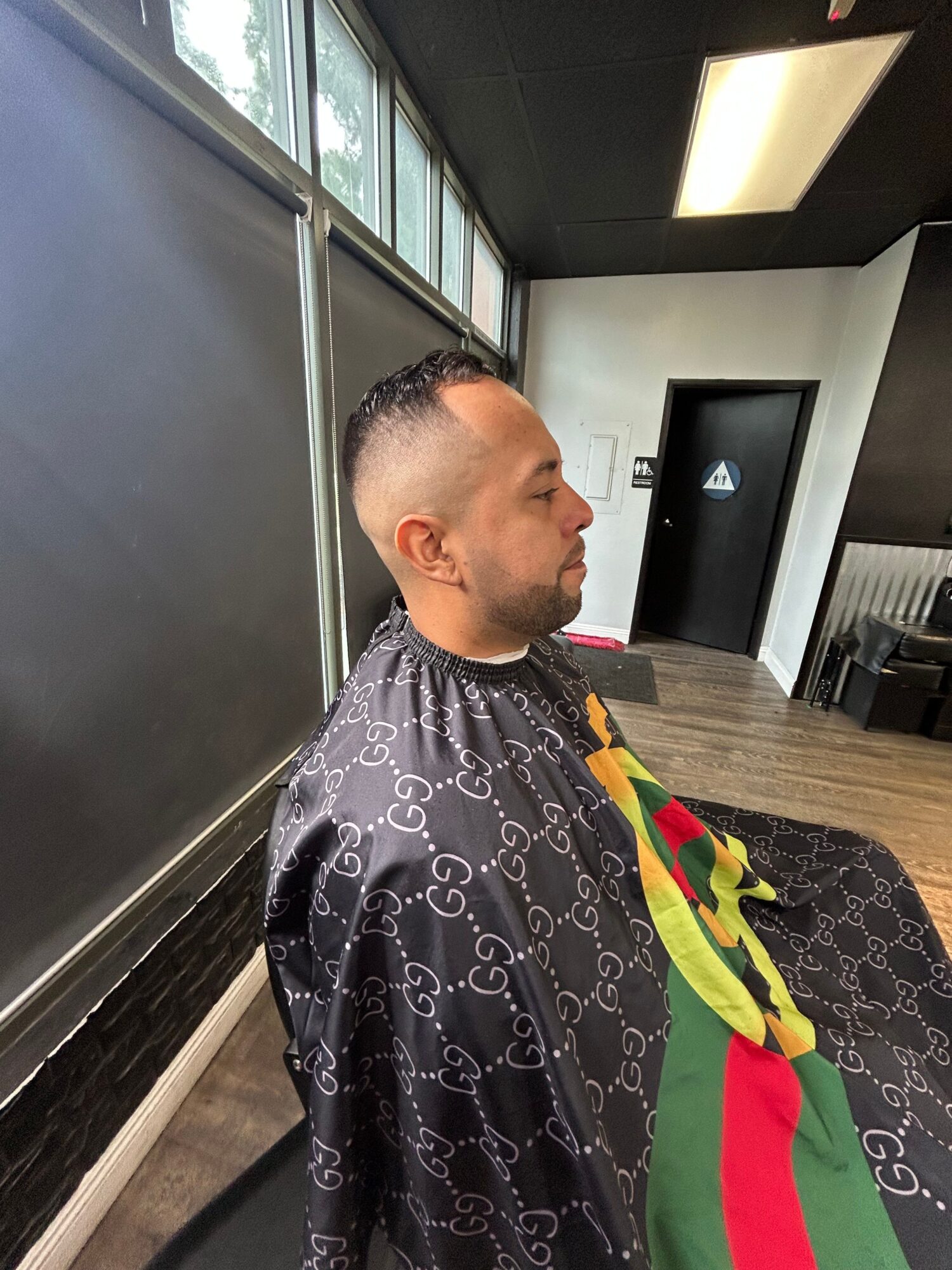 Person with short hair in a barber shop chair, wearing a black cape with a colorful stripe, facing left, with a mirror and window behind.