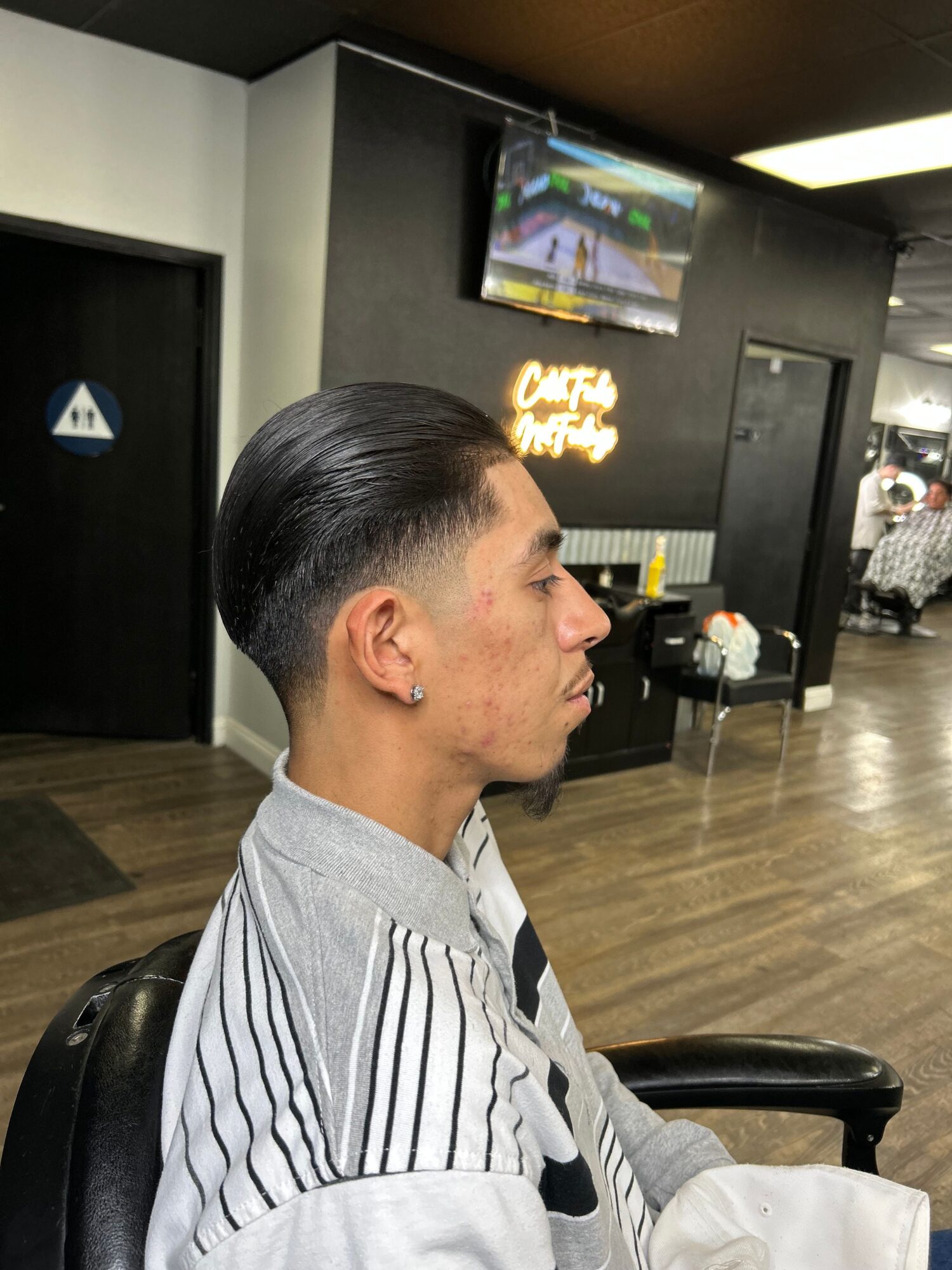 Side view of a man with slicked-back hair sitting in a barber shop chair, looking to the left, with a black wall and TV in background.