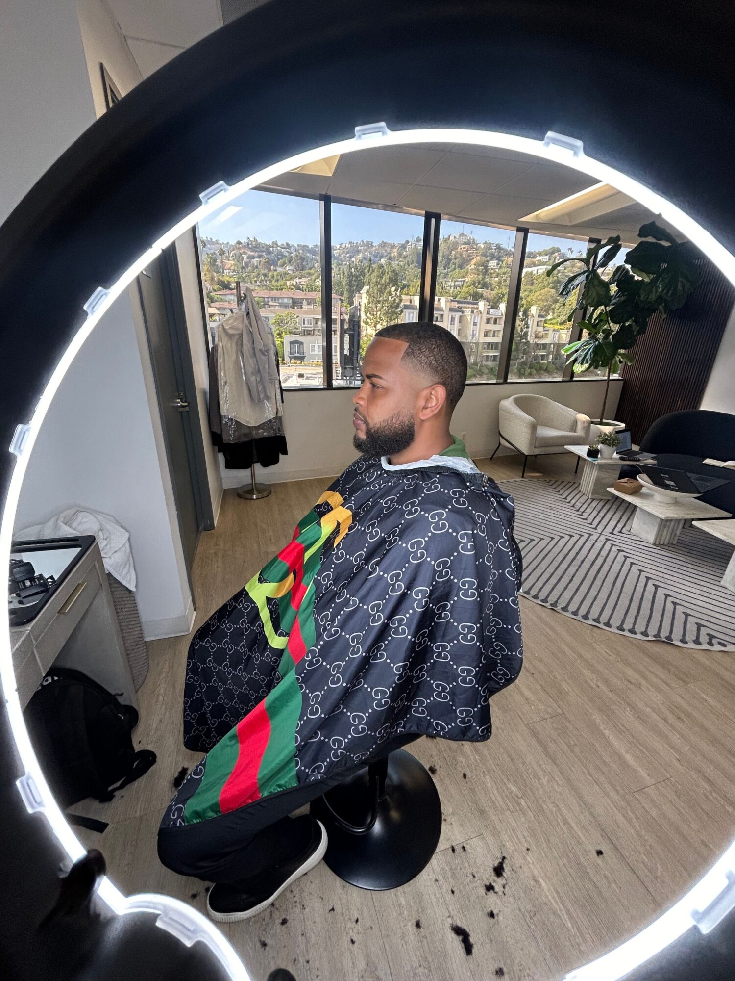 Person with short hair and patterned jacket sitting on a black scooter inside a room with large windows and wooden flooring.