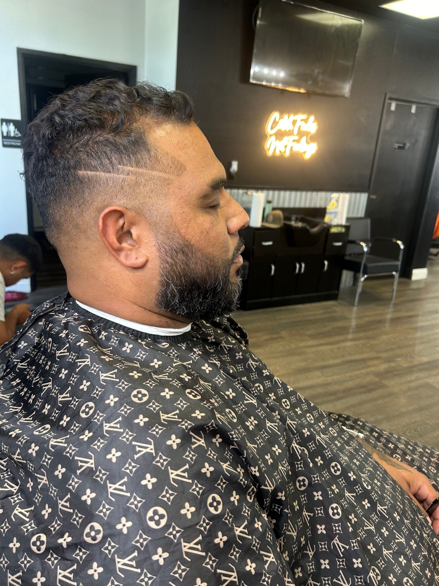Man with a patterned shirt and a fade haircut sitting in a barbershop, facing left, with salon equipment in the background.