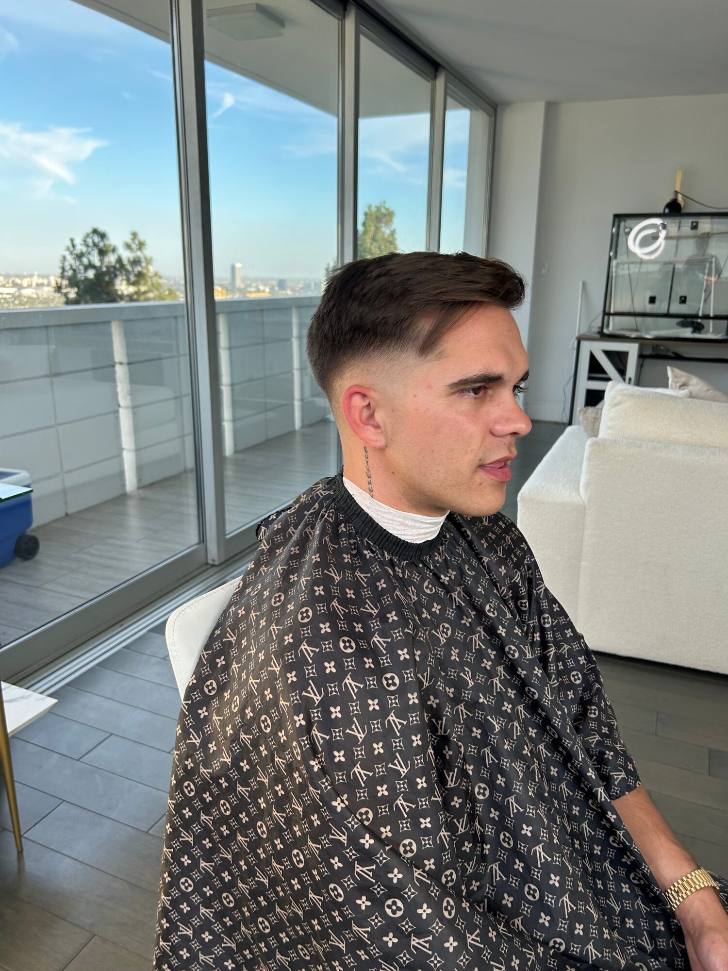 Young man with styled hair and earrings, wearing a patterned cape, looking out a large window with a city view.