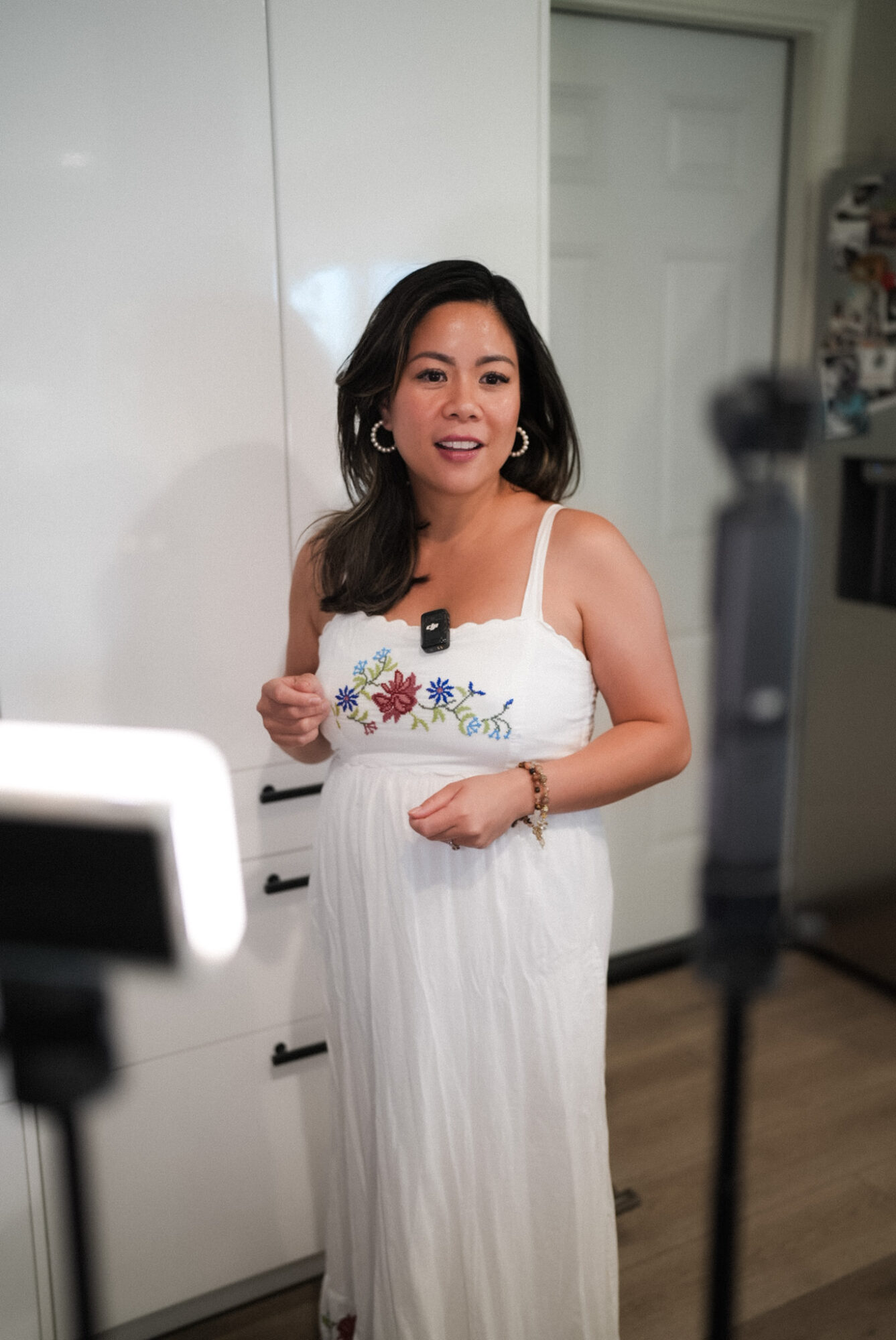 Woman in white dress standing indoors, smiling, with a white cabinet and a door in the background.