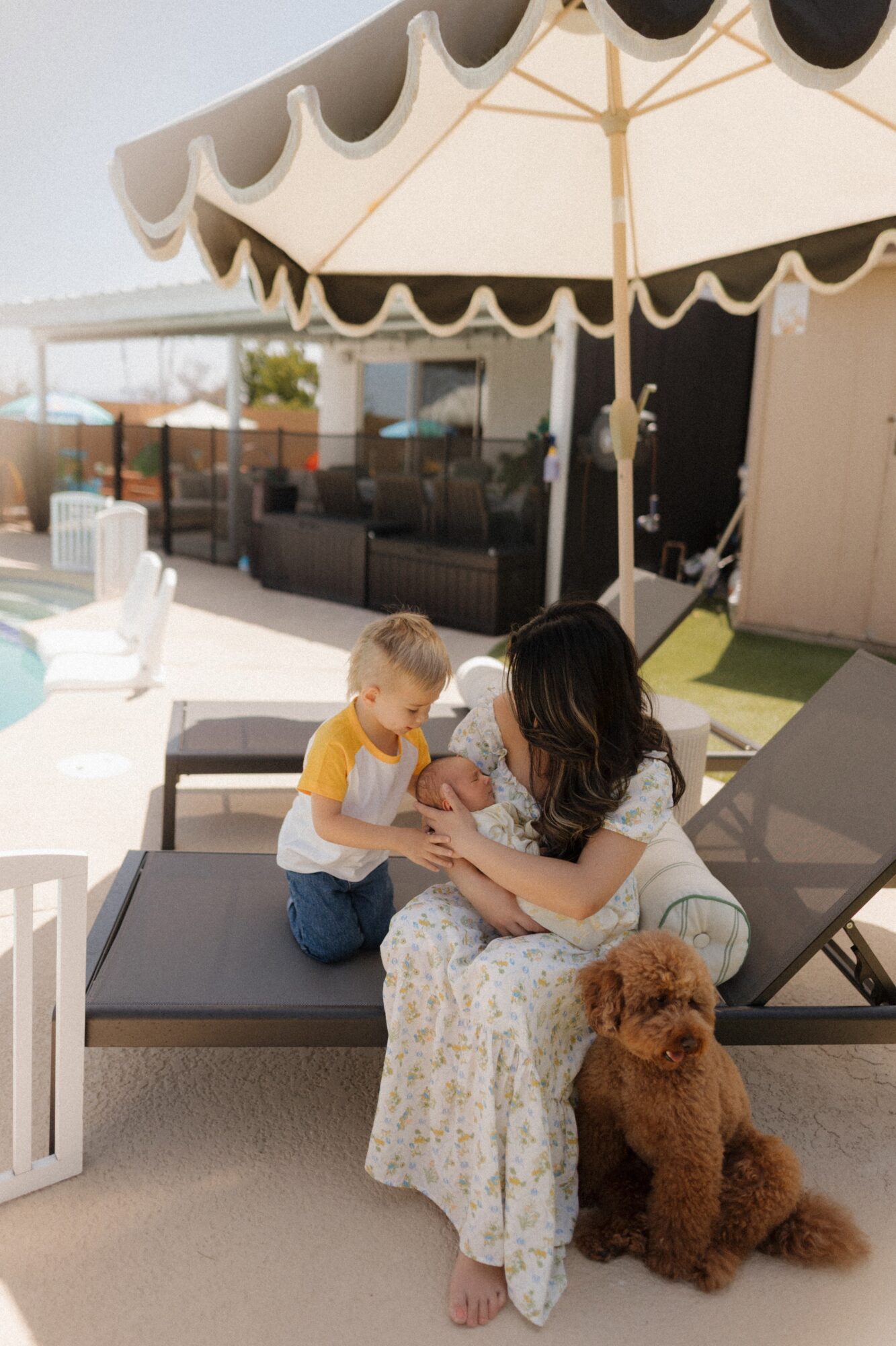 Woman and child sitting on lounge chair under umbrella, with a brown dog nearby, in a backyard setting.