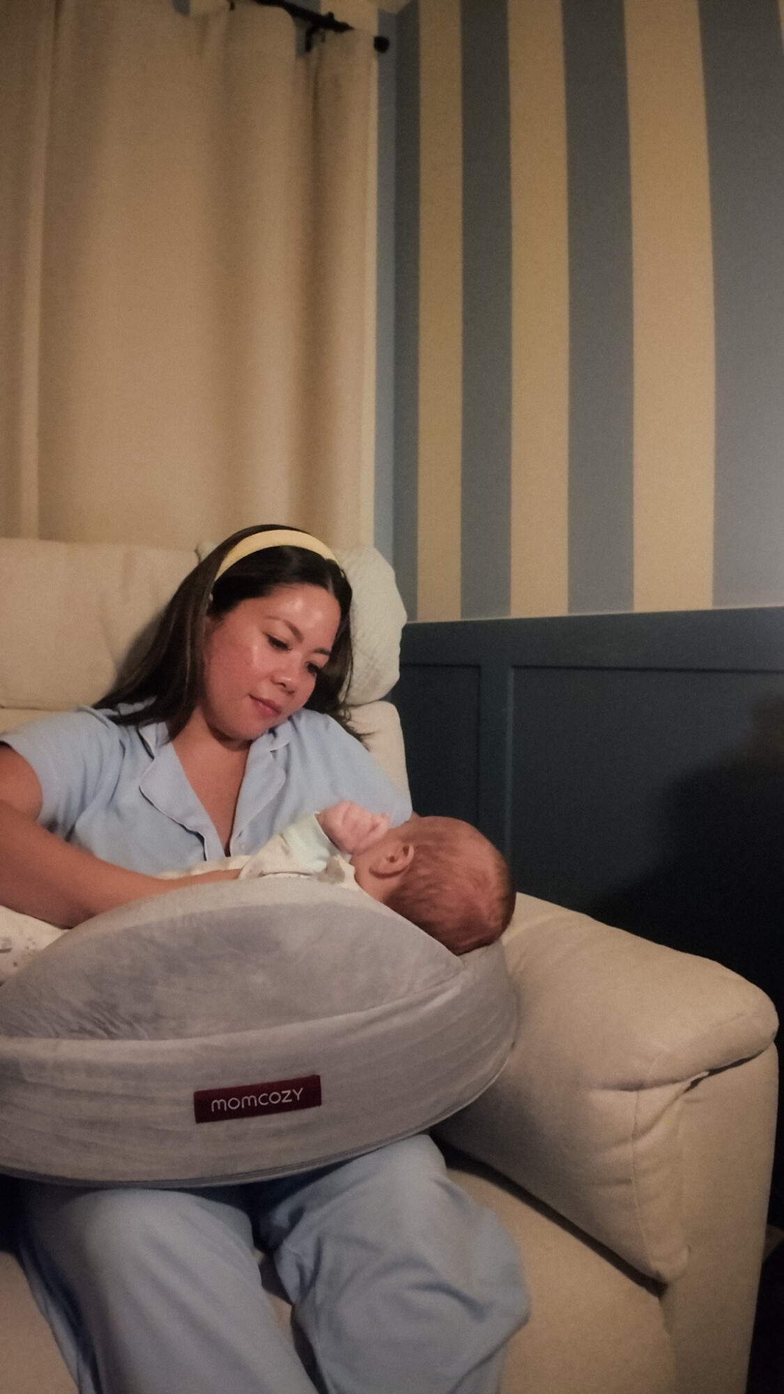 Woman holding a newborn baby in a hospital bed, with striped wallpaper and curtains in the background.