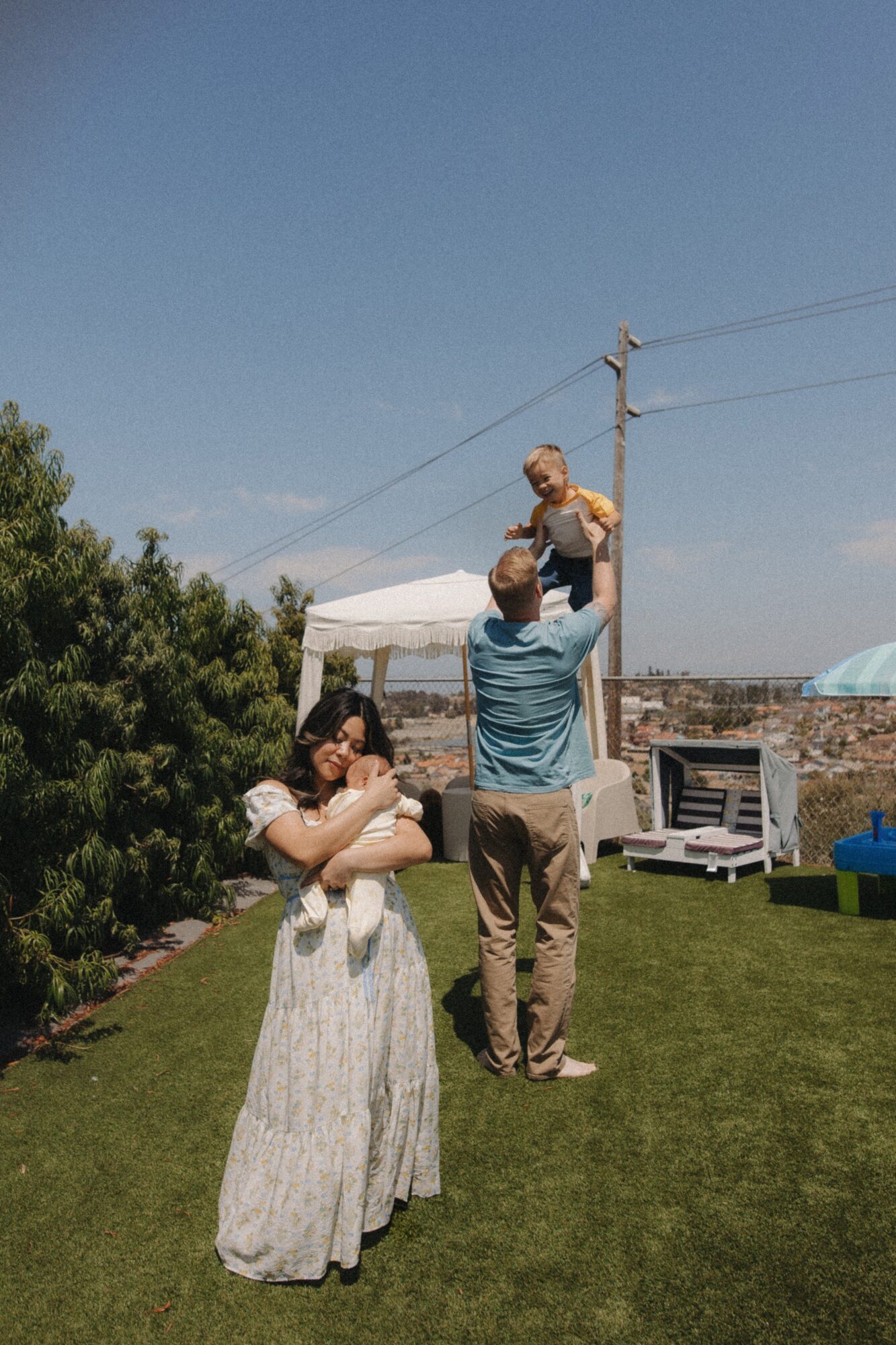Group of people outdoors on a sunny day, with a man lifting a child, a woman holding a baby, and a blue sky.