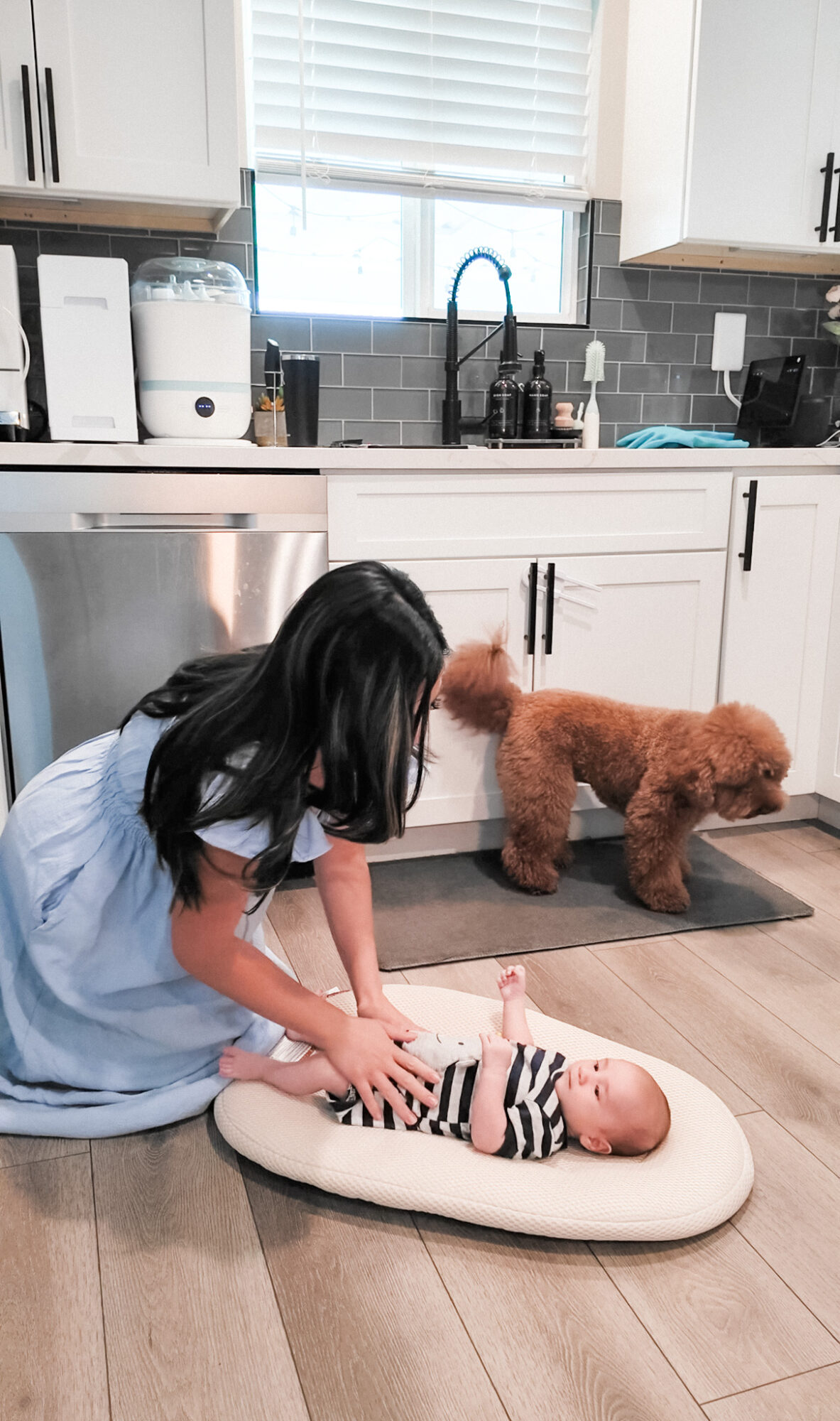 Child in blue dress leaning over a baby on a cushioned mat, dog nearby in kitchen with white cabinets and window.