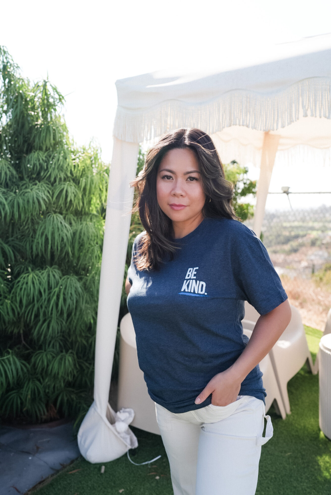 Woman with dark hair wearing a blue t-shirt and white pants standing outdoors near a white canopy, greenery in background.
