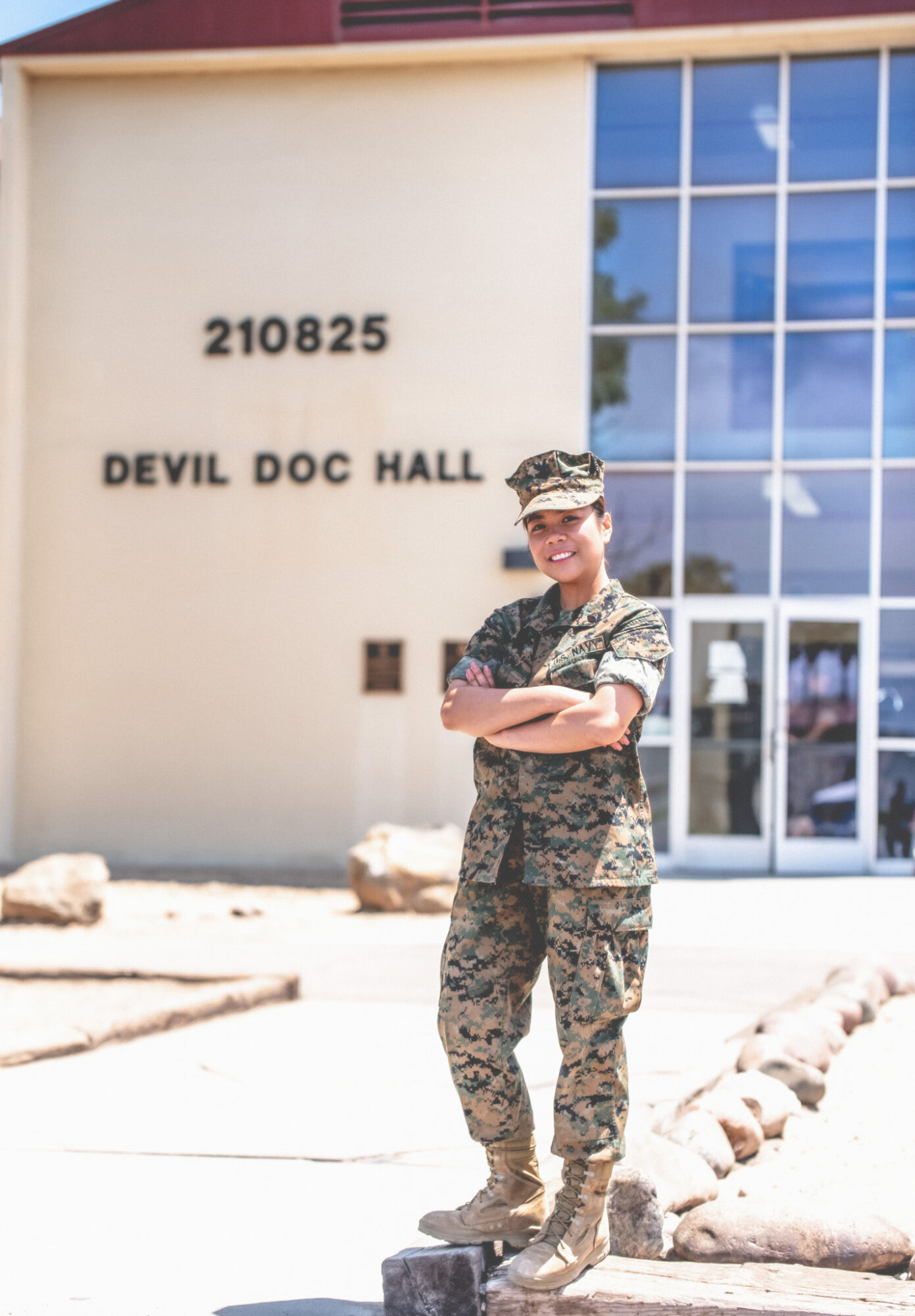Person in military uniform standing outside building with glass windows, arms crossed, smiling, in front of sign reading 210825 DEVIL DOC HALL.