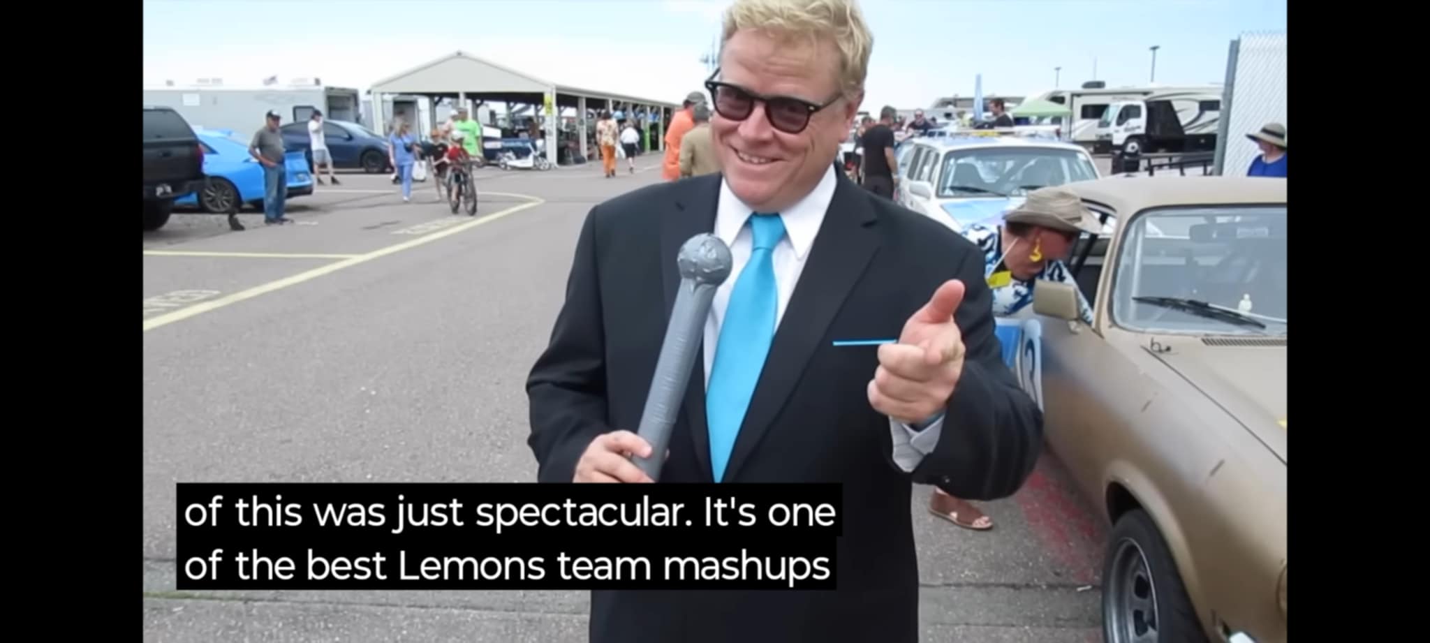 Man in suit holding microphone, speaking outdoors, with cars and tents in background, smiling and pointing, subtitle about Lemons team mashups.