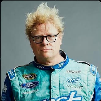 Portrait of a man with messy blonde hair, glasses, wearing a racing suit with logos, against a plain background.
