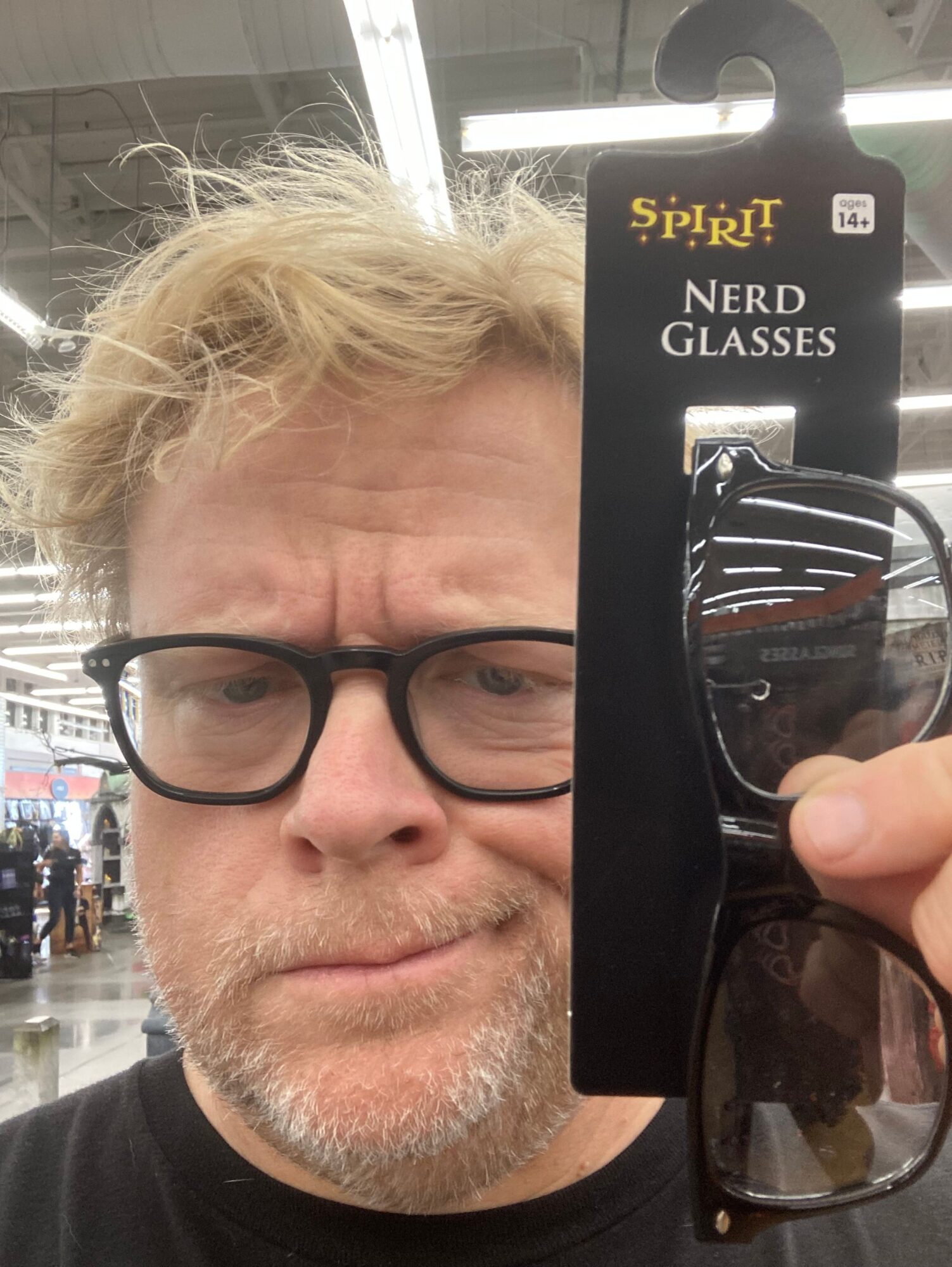 Person with glasses holding sunglasses in front of face, blonde hair, and a beard, taking a selfie in a store.
