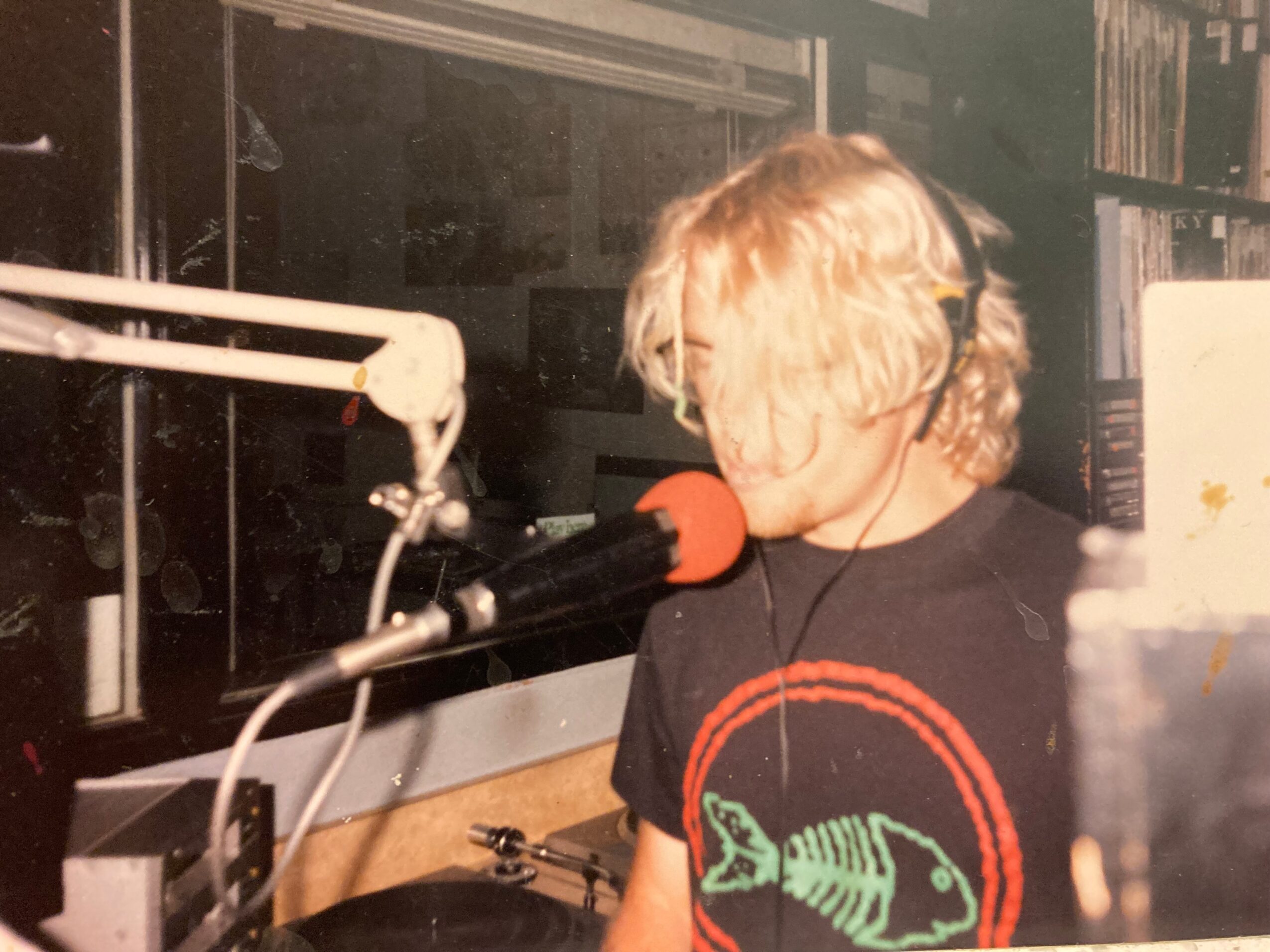 Child with blonde hair wearing headphones, speaking into a microphone in a recording studio, with equipment and books in background.