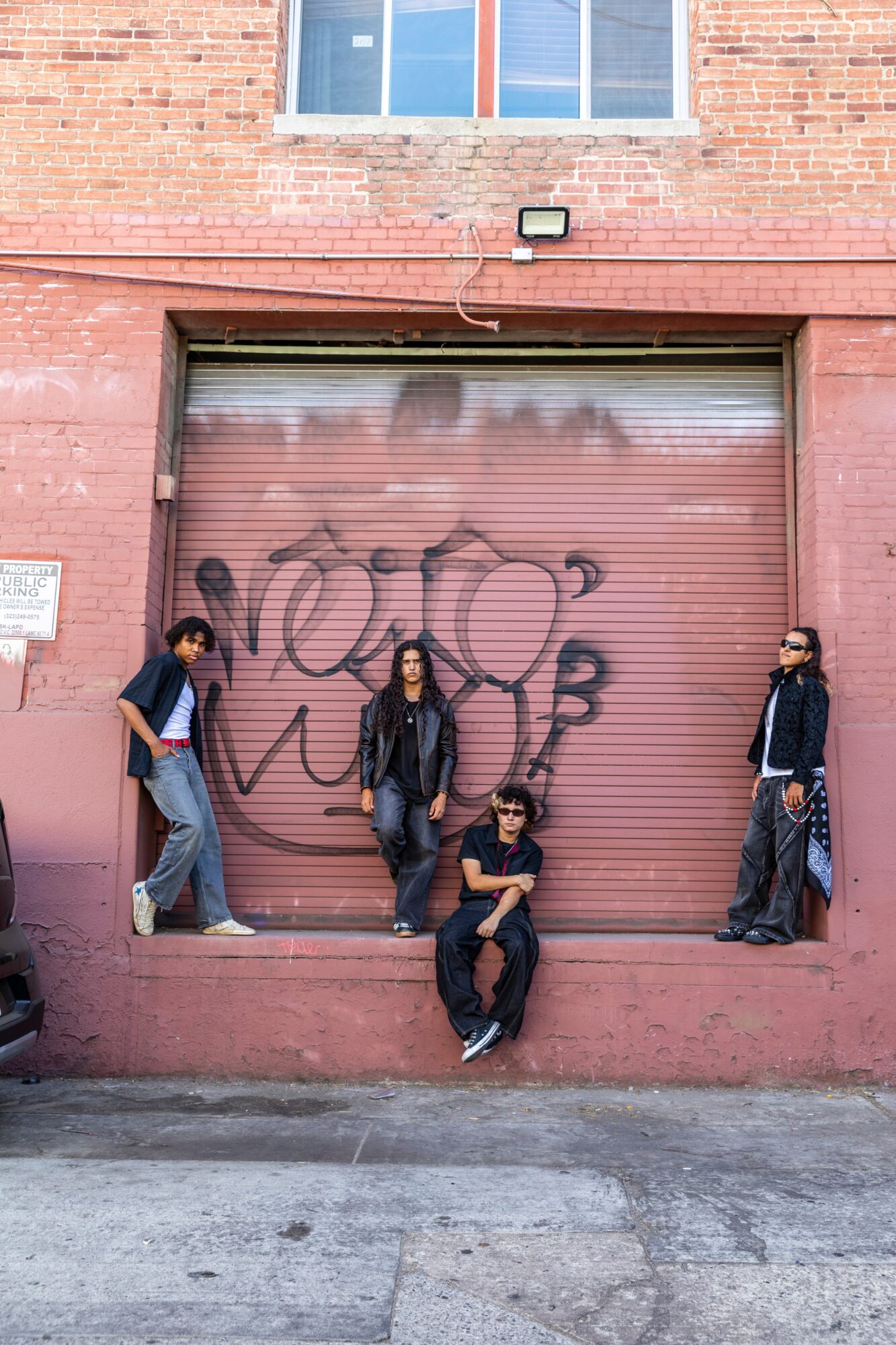 Four people pose in front of a pink garage door with graffiti, in an urban setting with a brick building above.
