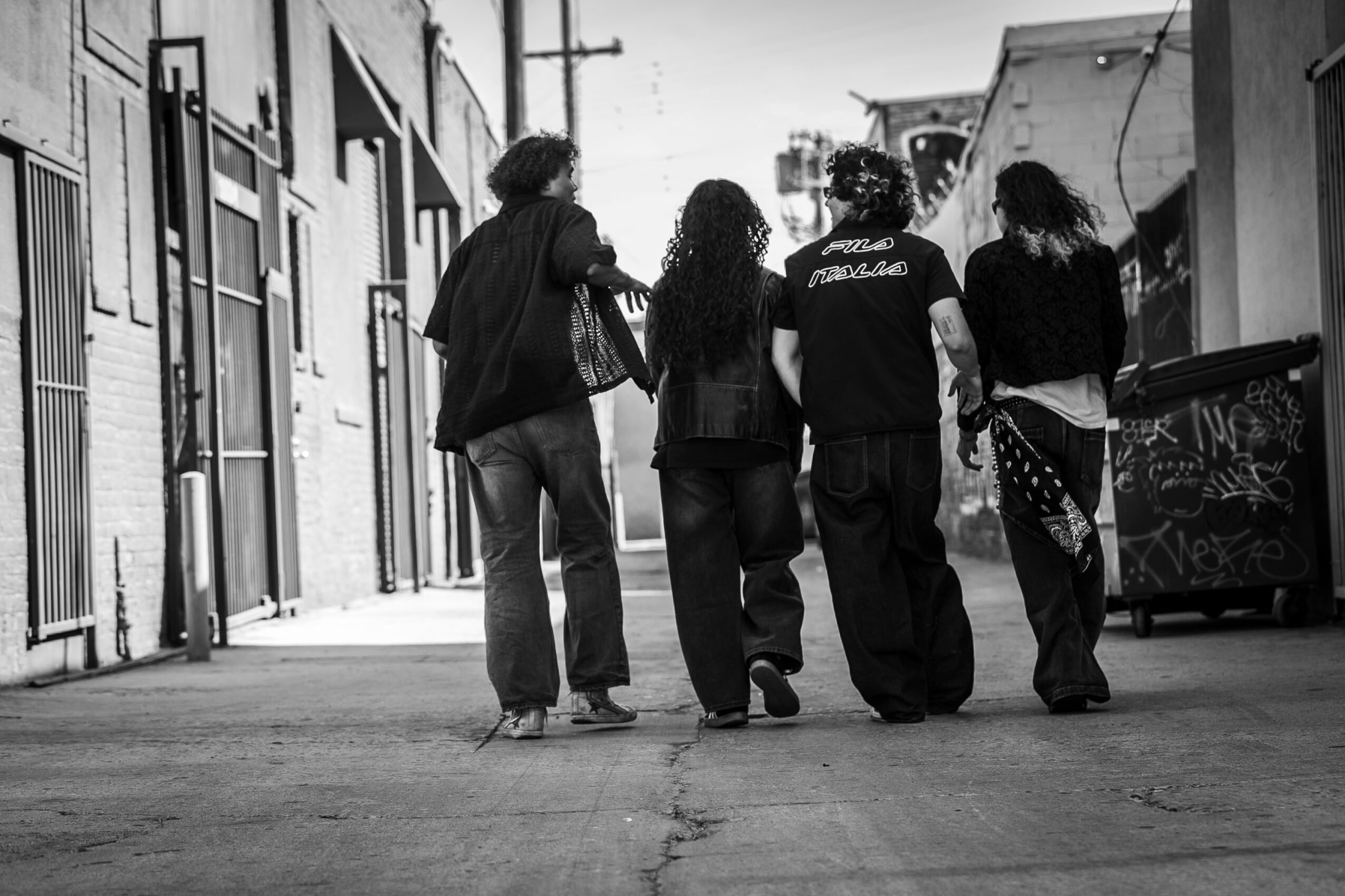 Four people walk down an urban alleyway, with buildings and a trash bin visible, in black and white.