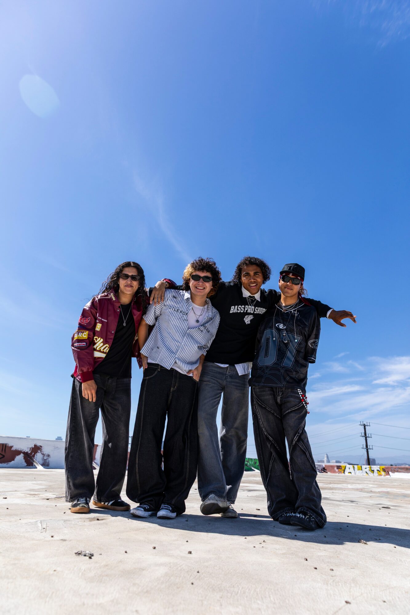 Four people standing outdoors on a sunny day, smiling, with a clear blue sky above and a flat landscape behind.