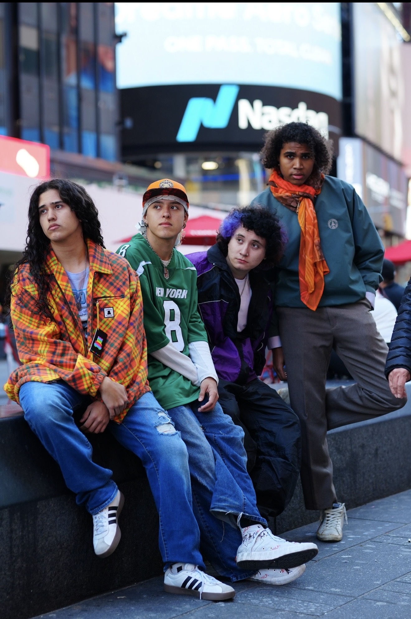 Four young people sitting on a ledge in an urban area with a large digital billboard behind them, wearing colorful casual clothing.
