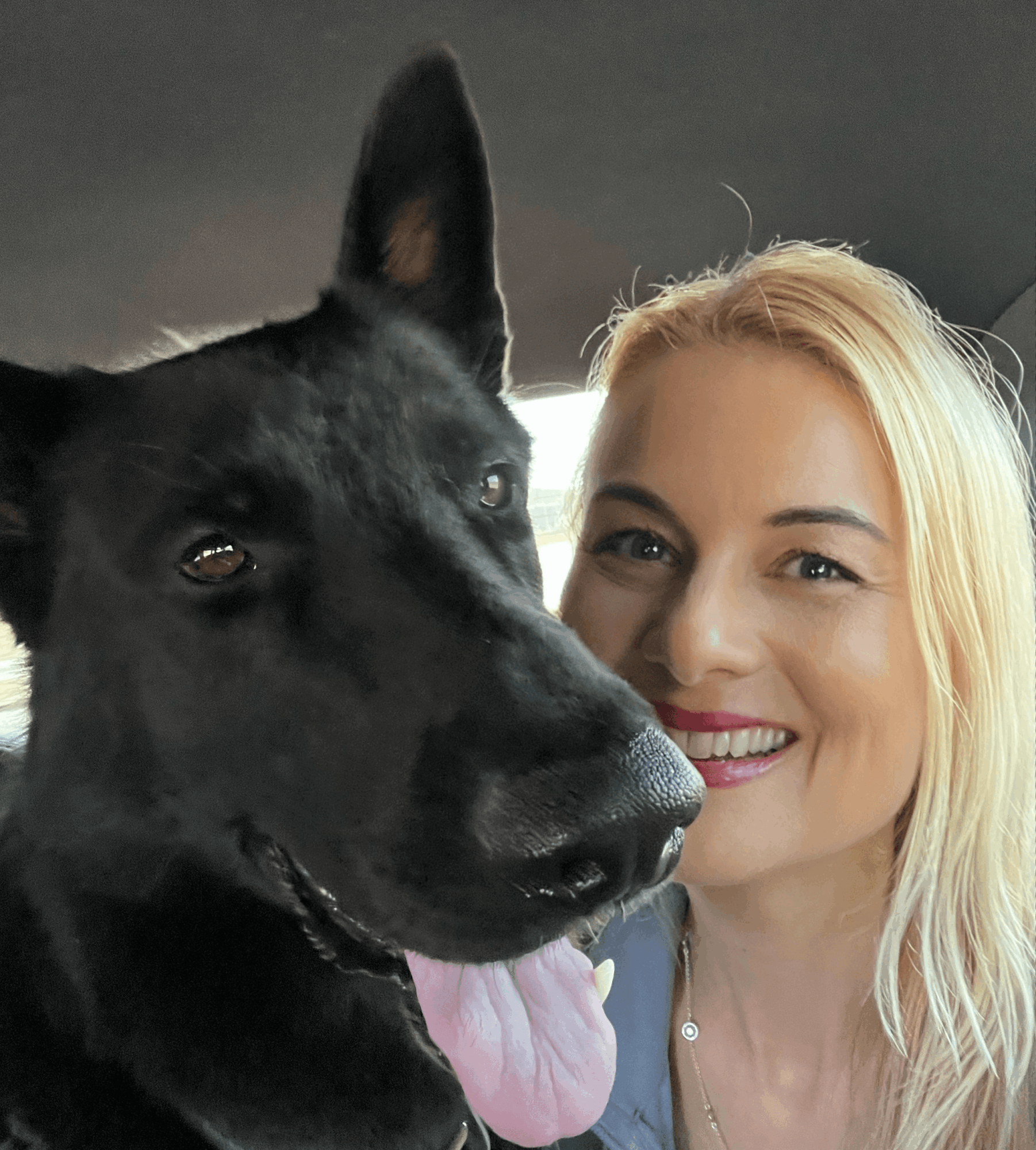 Woman smiling with a black dog, both close to the camera, indoors, woman has blonde hair and is wearing a necklace.