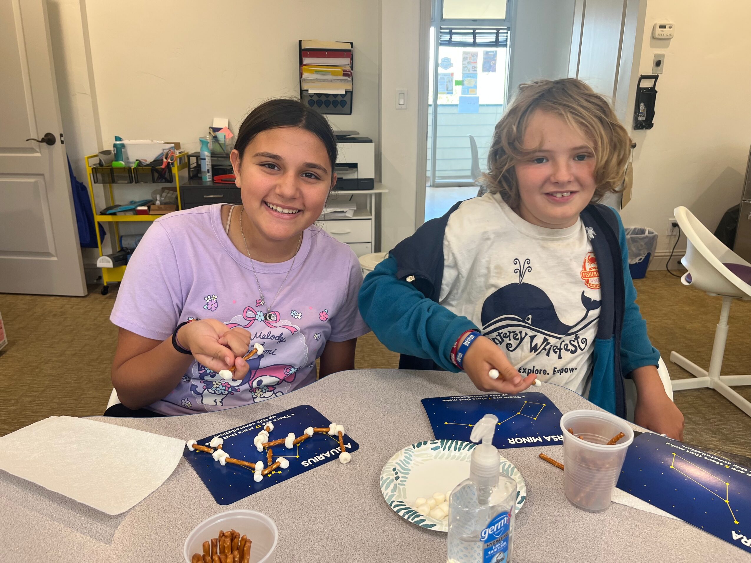 Two smiling children sit at a table with craft supplies, in a room with a door and shelves in the background.