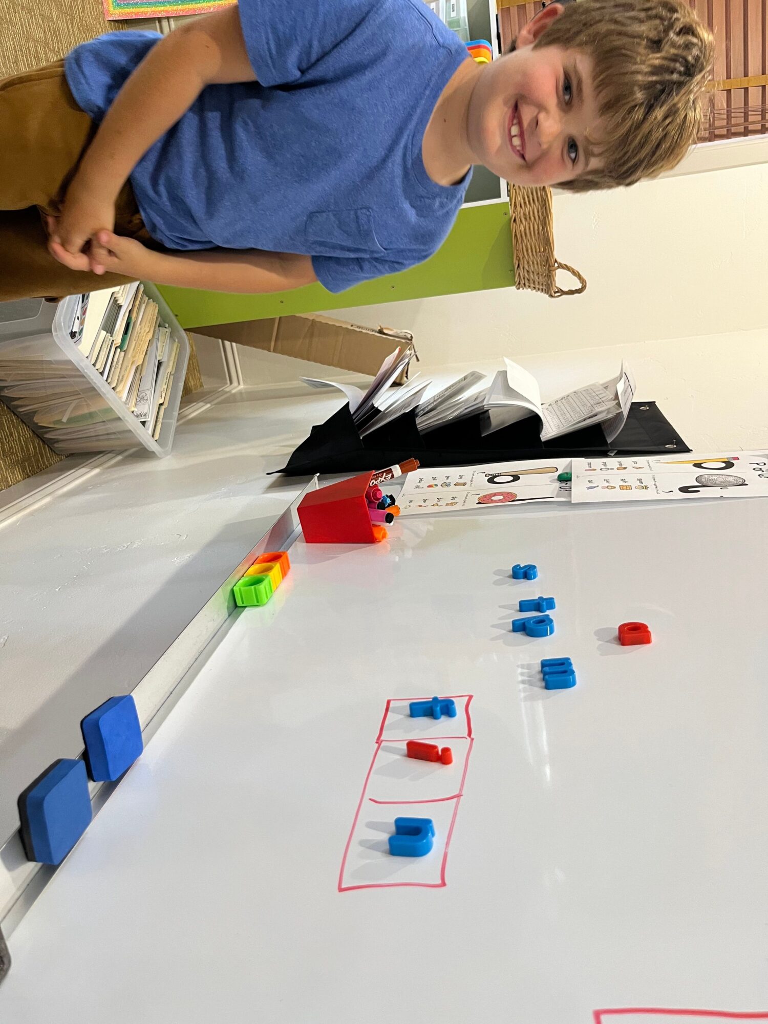 Child smiling in a classroom with a whiteboard and magnetic markers, wearing a blue shirt.