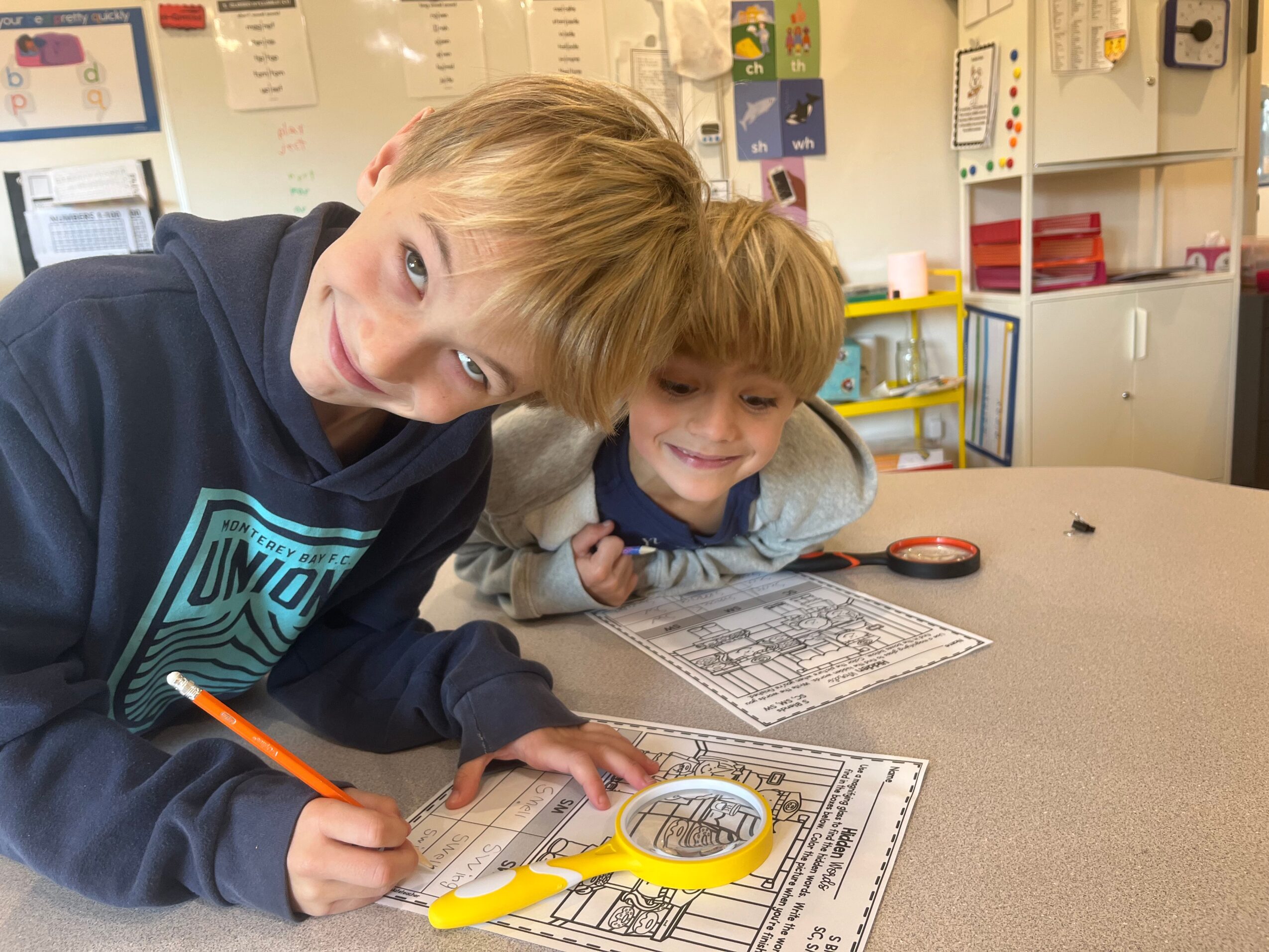 Two children smiling and working on a worksheet at a table in a classroom.