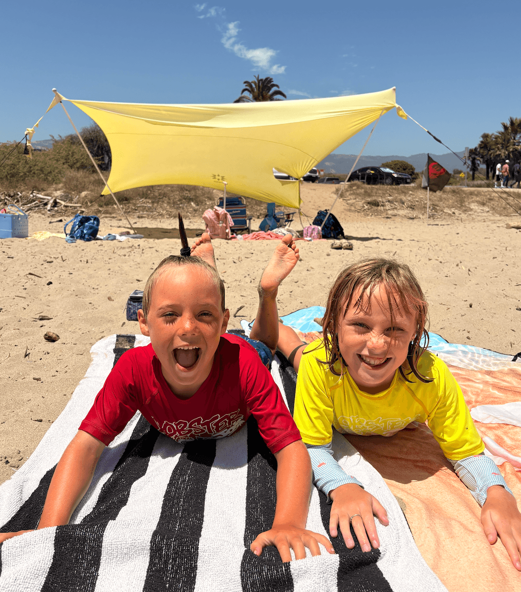 Two children lying on towels on the beach, smiling and waving, with a yellow shade sail and palm trees in background.