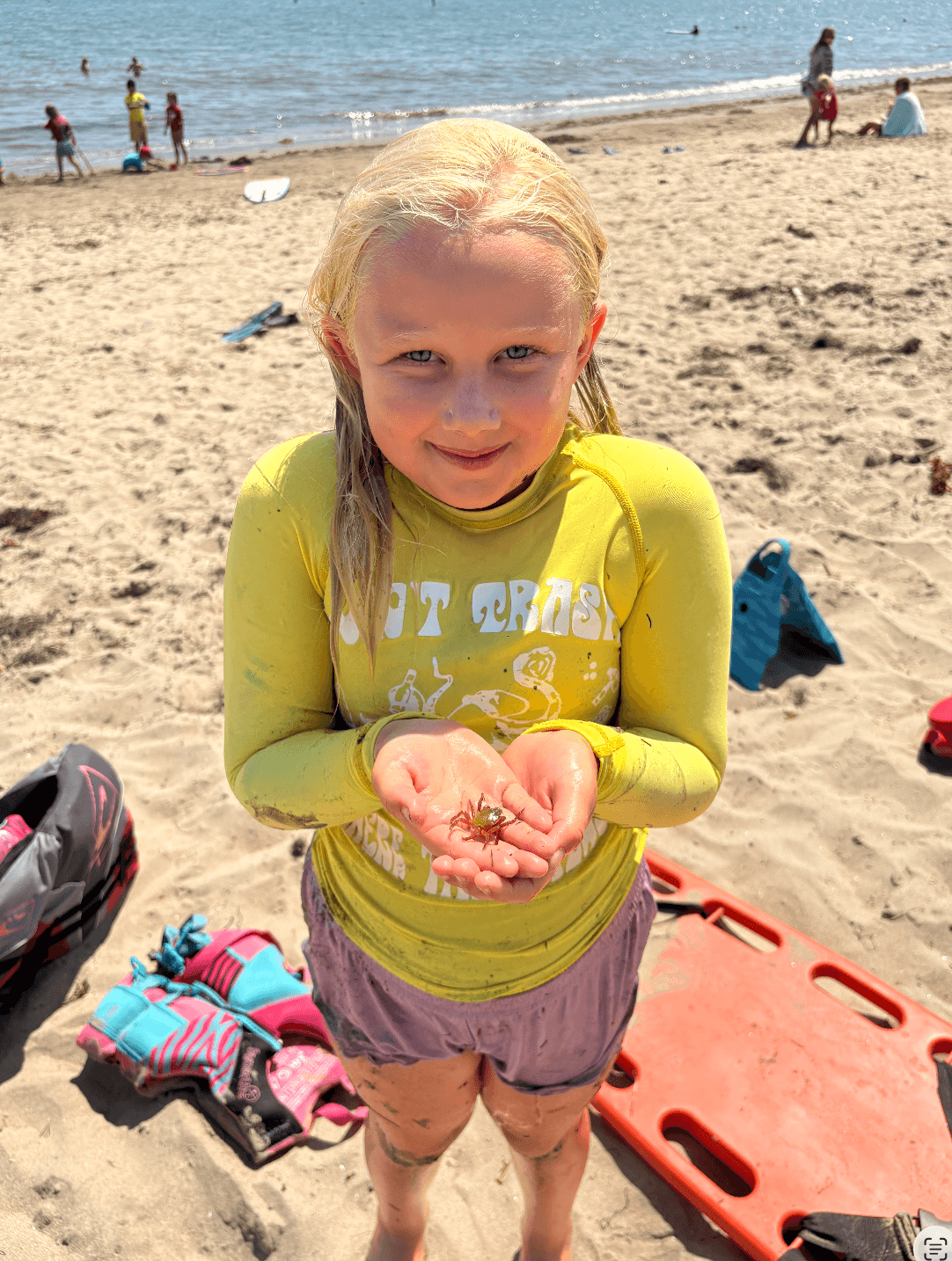 Young girl with blonde hair smiling at the camera, holding sand on a beach with ocean and people in the background.