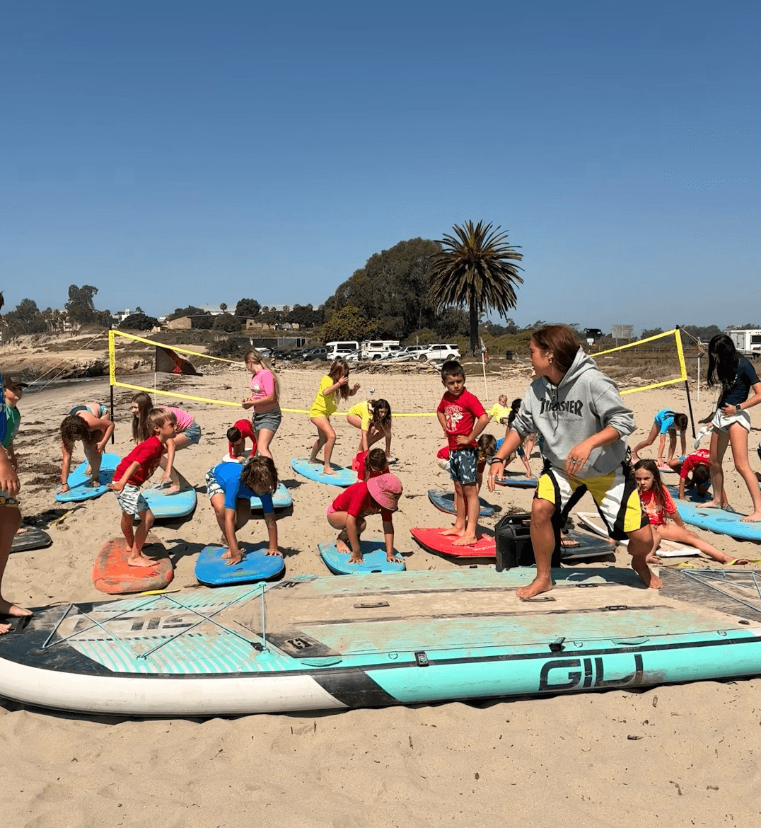 Group of children and an instructor on a sandy beach, practicing paddleboarding with surfboards, under a clear blue sky.