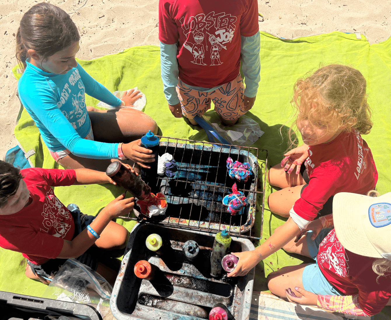 Four children with colorful hair and clothing are gathered around a small grill with painted eggs, outdoors on a yellow cloth.