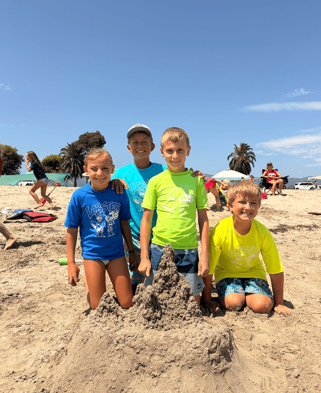 Four children at the beach, standing and kneeling around a sandcastle, with umbrellas and people in the background under a blue sky.