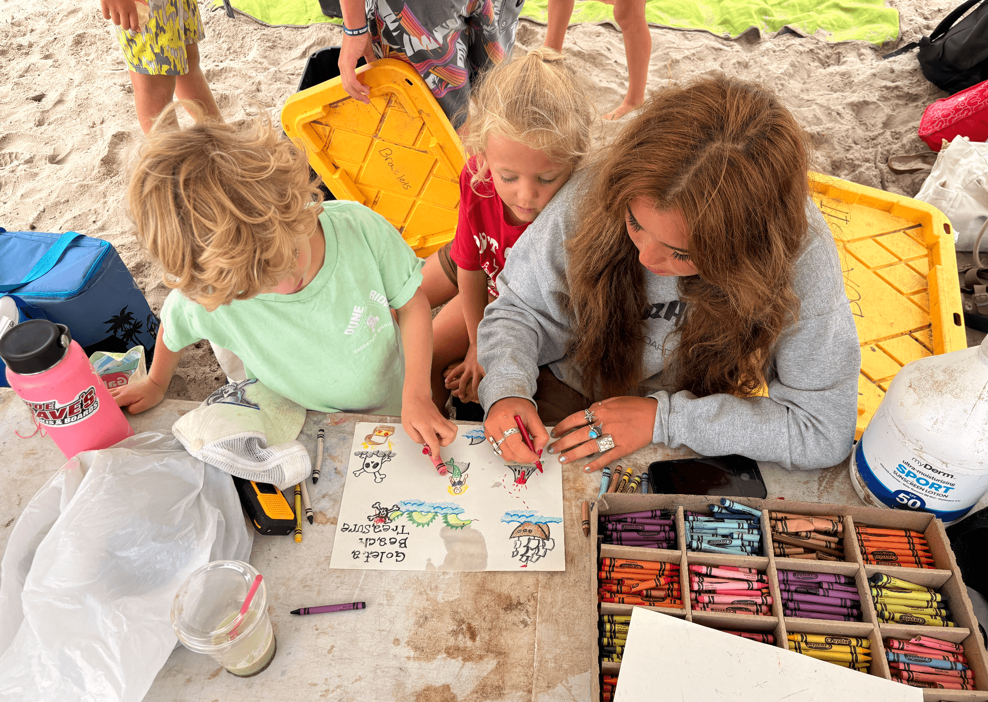 Children and an adult drawing and coloring at a table outdoors, with art supplies and a yellow container nearby.