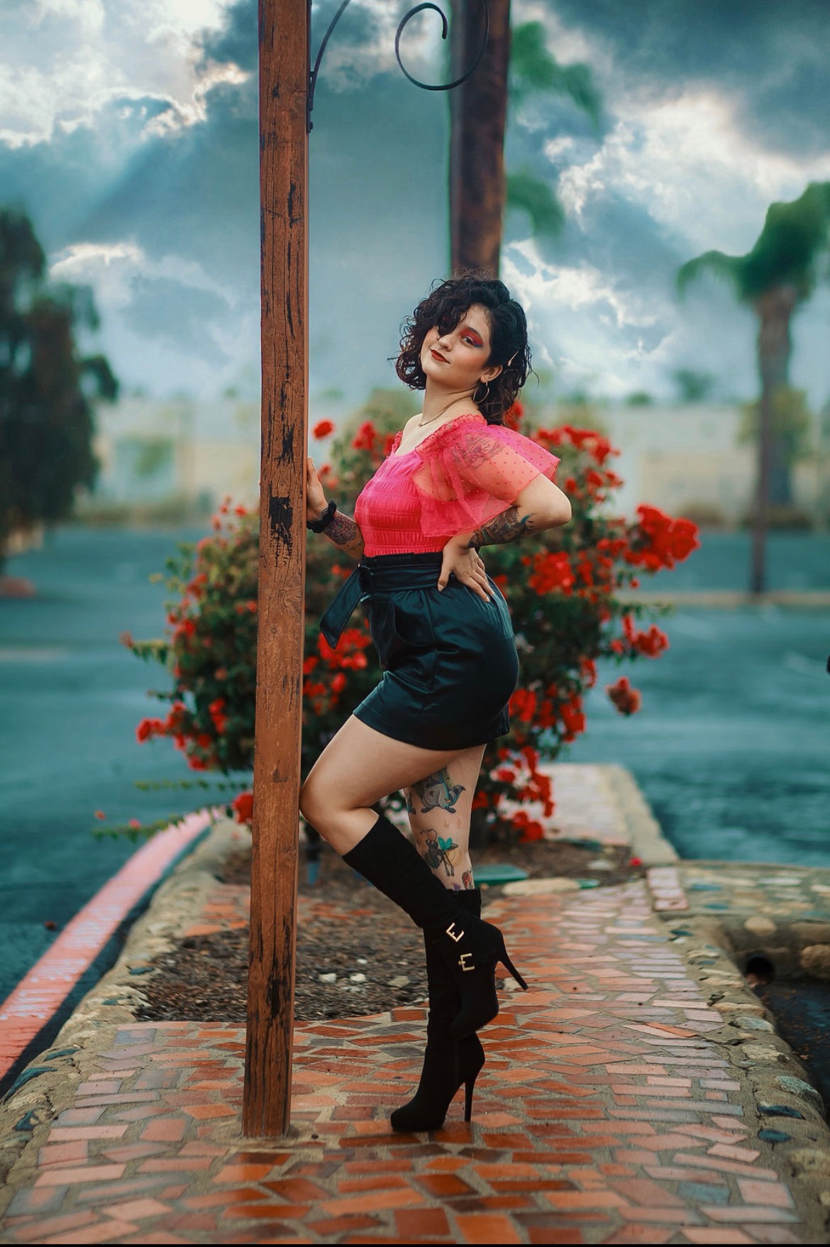 Woman in pink top and black shorts standing on brick pathway with flowers and trees in background.