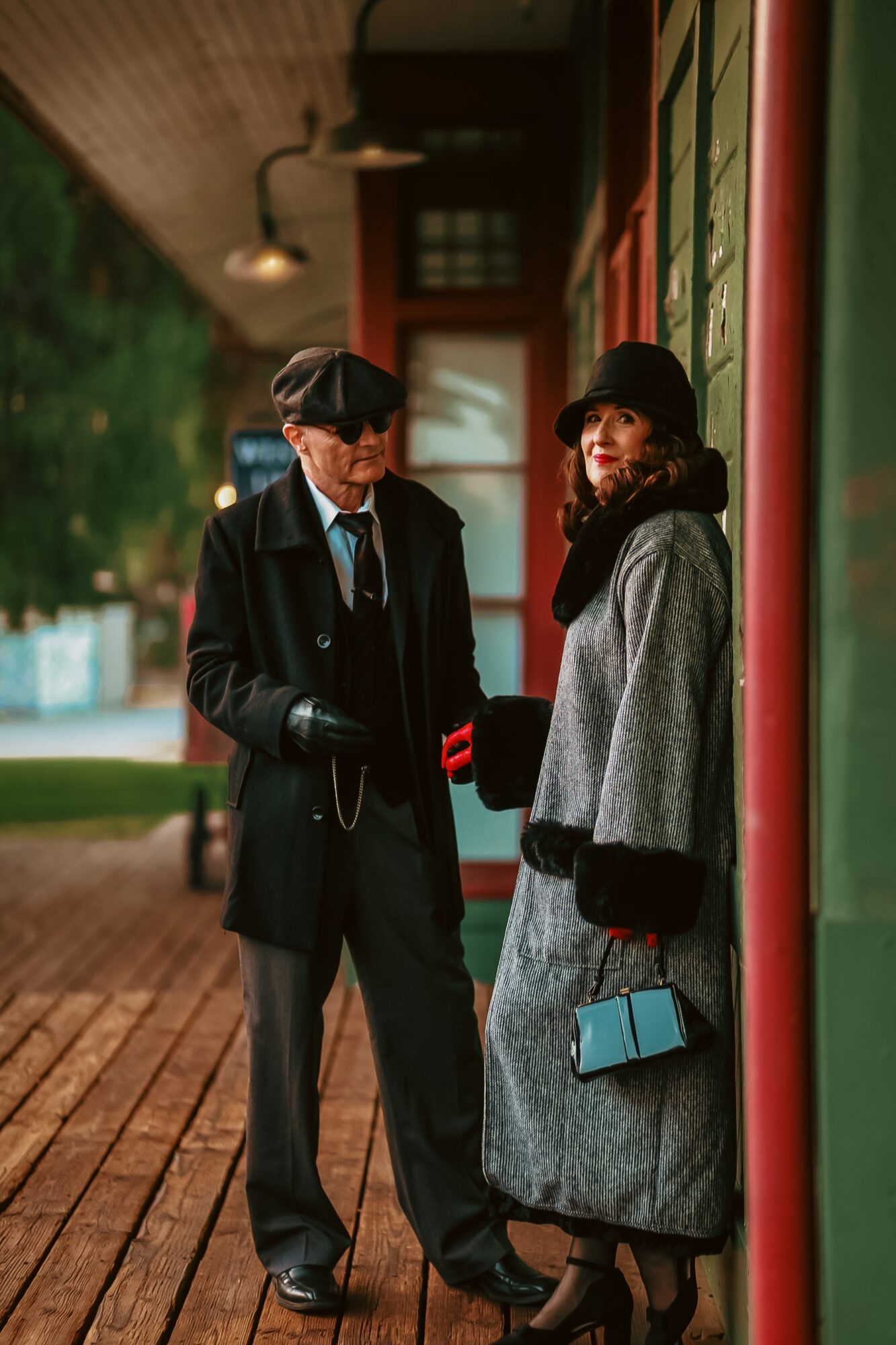 Two people in vintage clothing stand outside a building with green and red walls, talking under a covered porch.