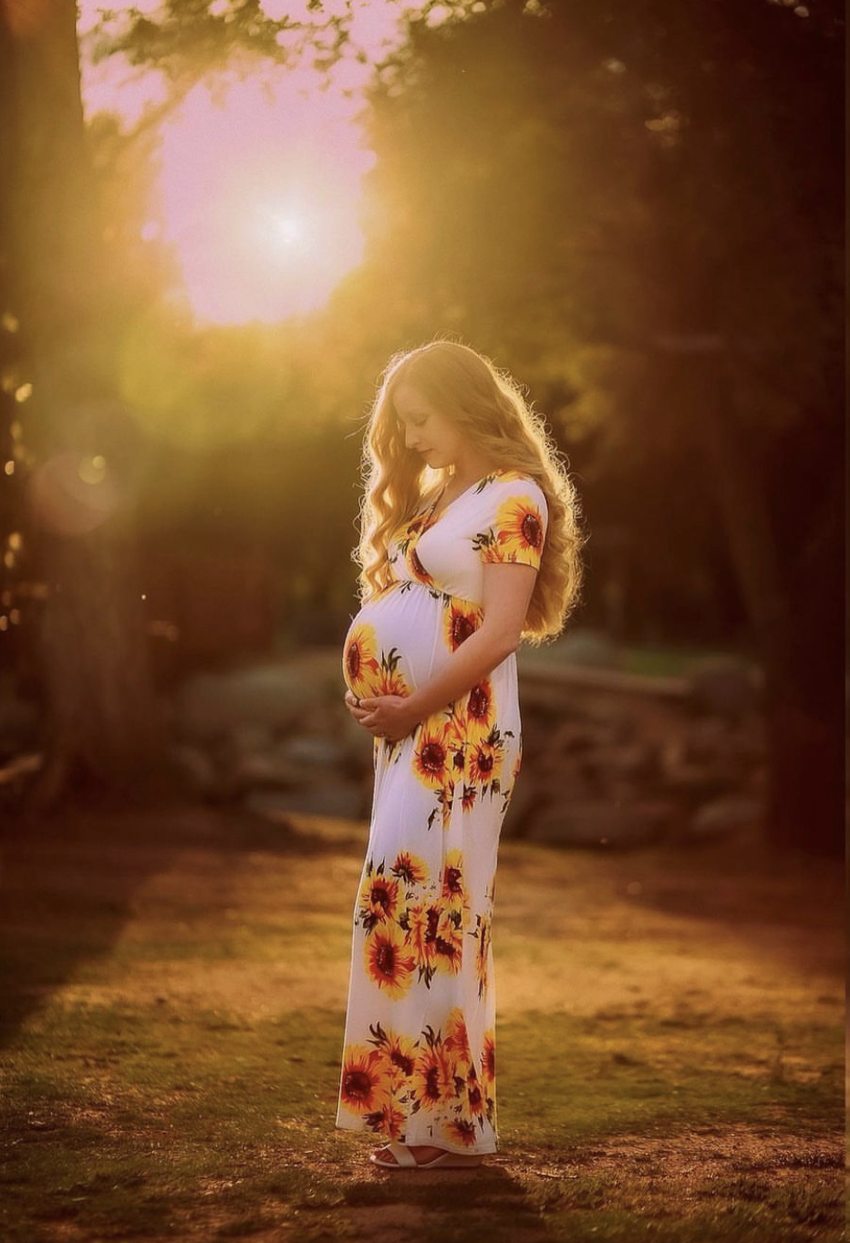 Pregnant woman in a floral dress standing outdoors at sunset, holding her belly, with sunlight shining behind her.