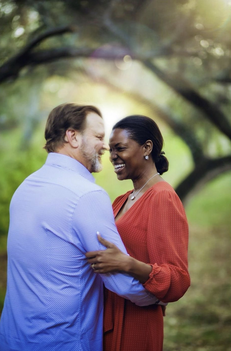 A smiling couple stands close together outdoors, embracing, with trees and sunlight in the background.
