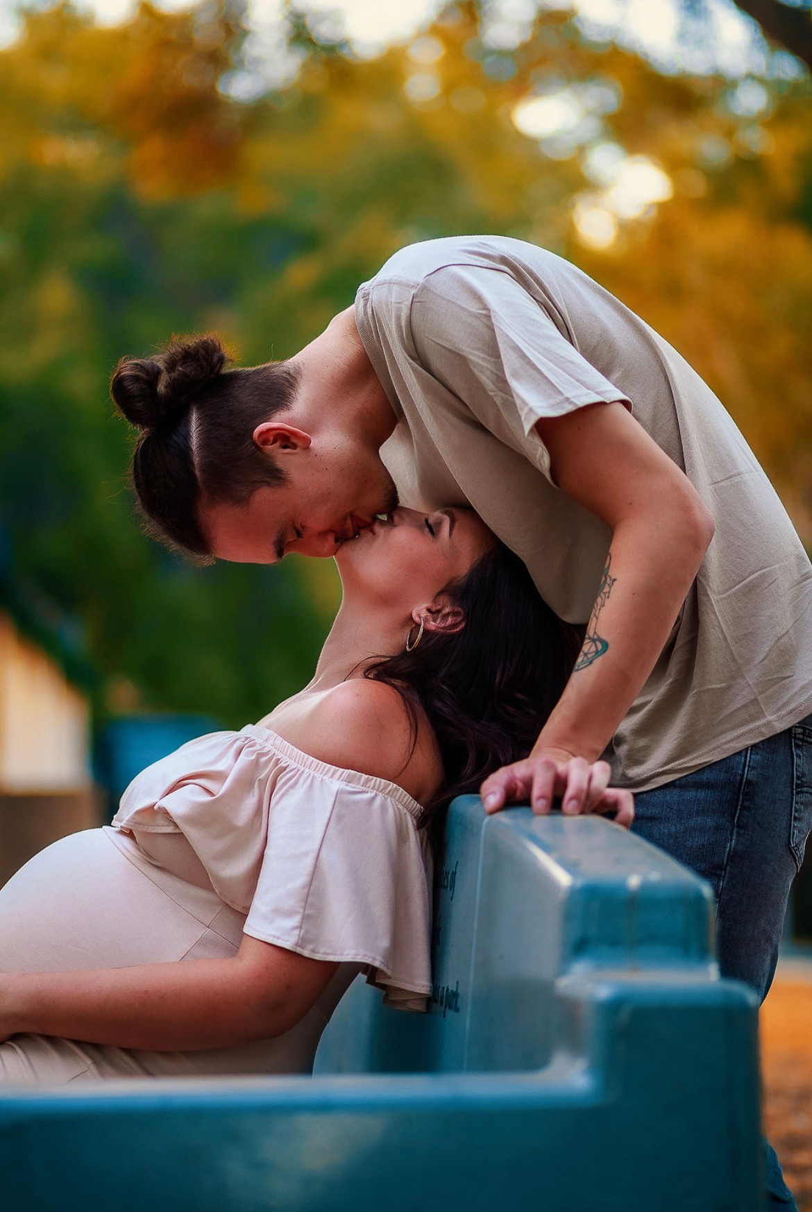 Two people share a kiss outdoors, with trees and sunlight in the background.