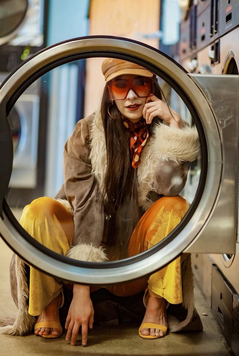 Woman with long dark hair, wearing sunglasses, a hat, and colorful clothing, crouches inside a washing machine door.