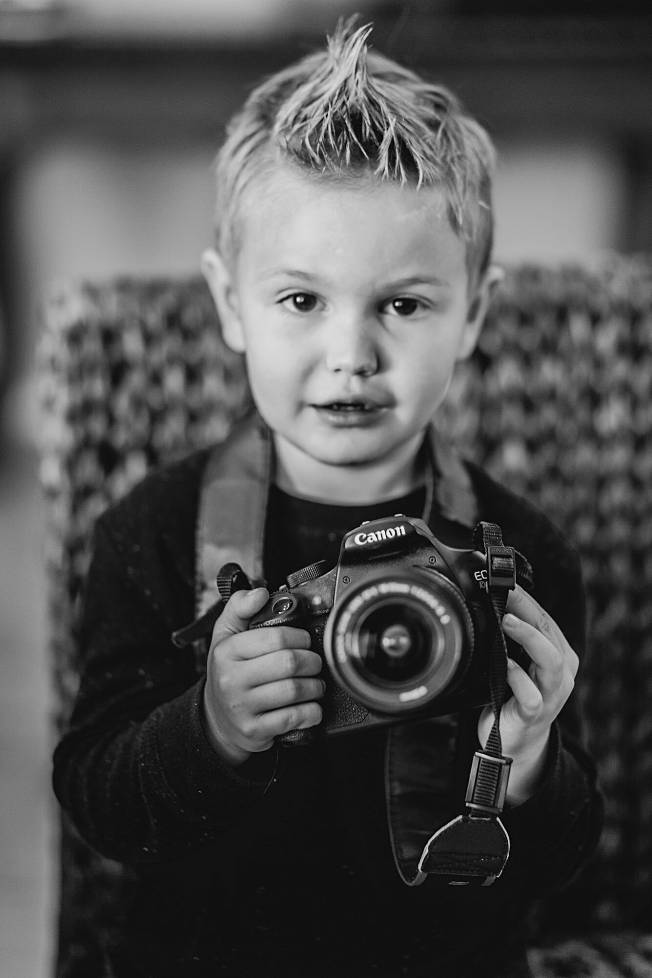 Young boy with spiky hair holding a camera, looking at the viewer, in black and white.