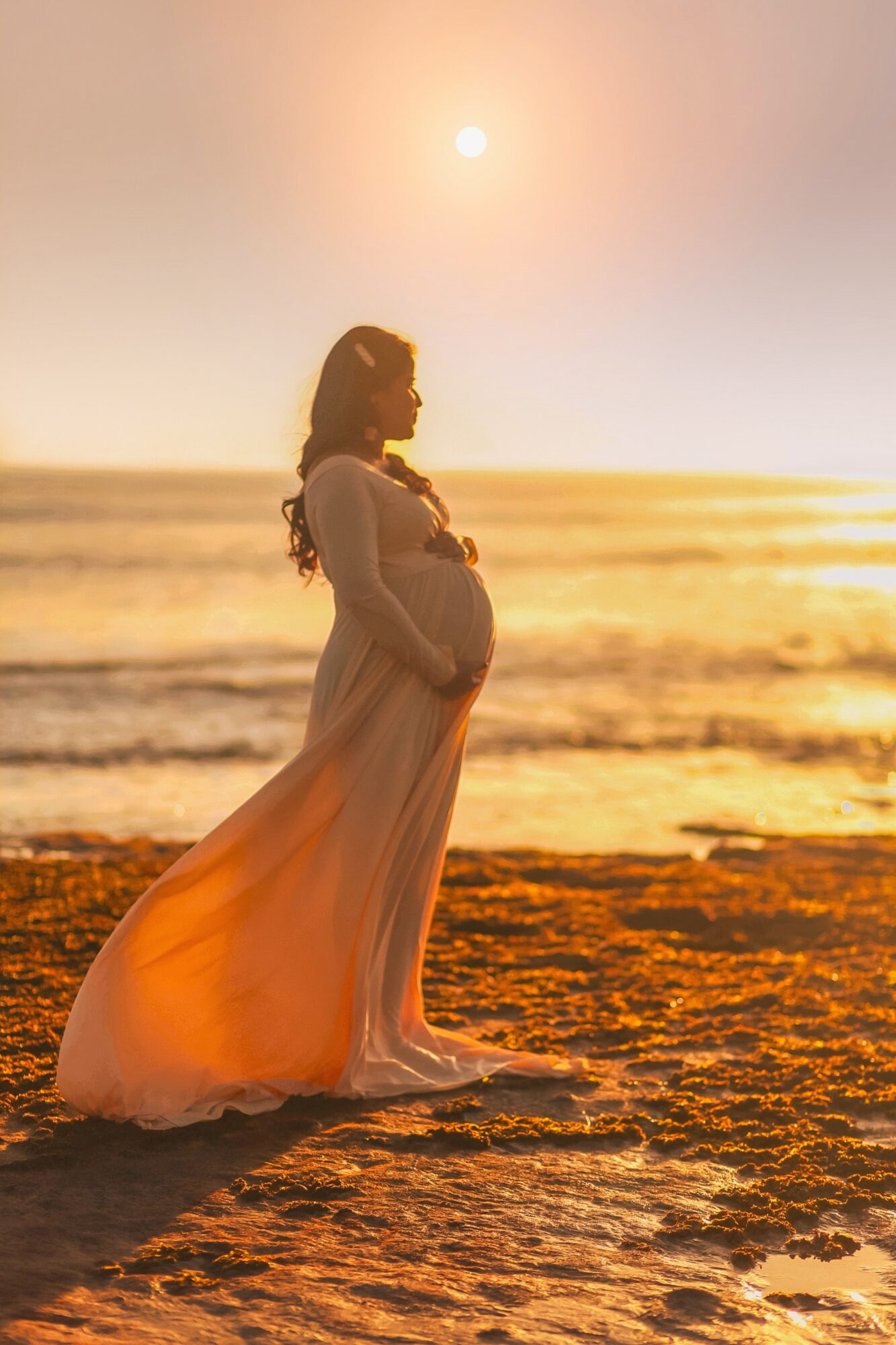 Pregnant woman in a flowing dress standing on the beach at sunset, facing the ocean with hands on her belly.