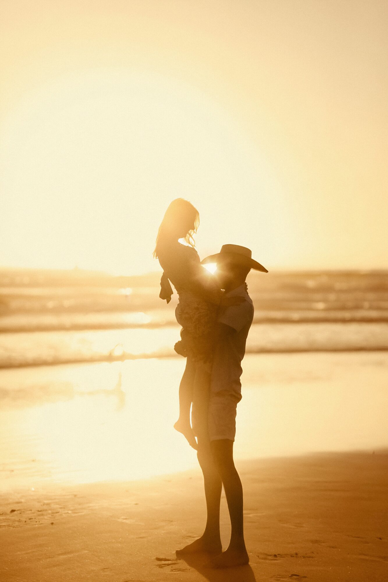 Person carrying a child on shoulders at the beach during sunset, with the sun low in the sky.
