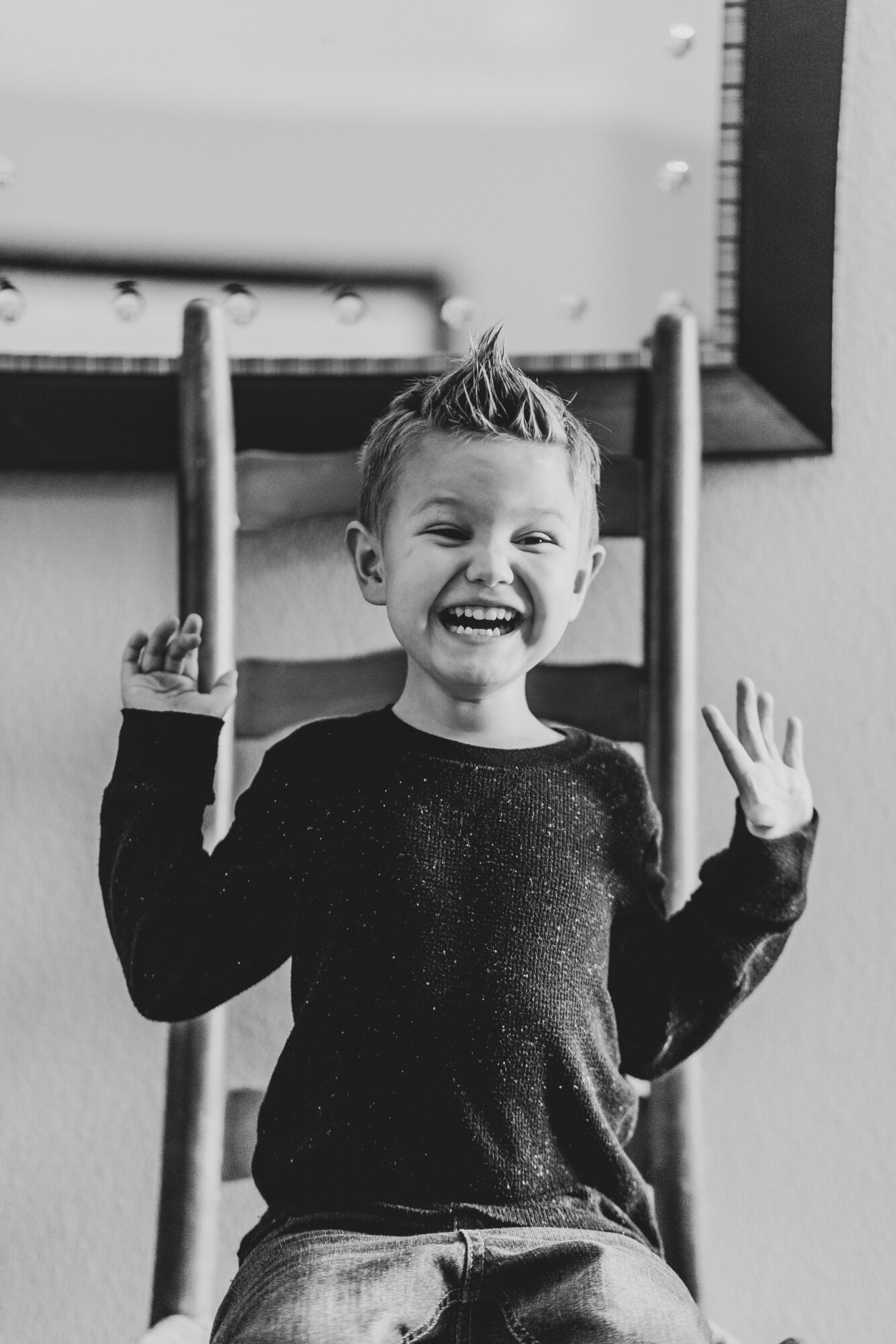 Child with short hair smiling and raising hands, standing in front of a ladder, in black and white.