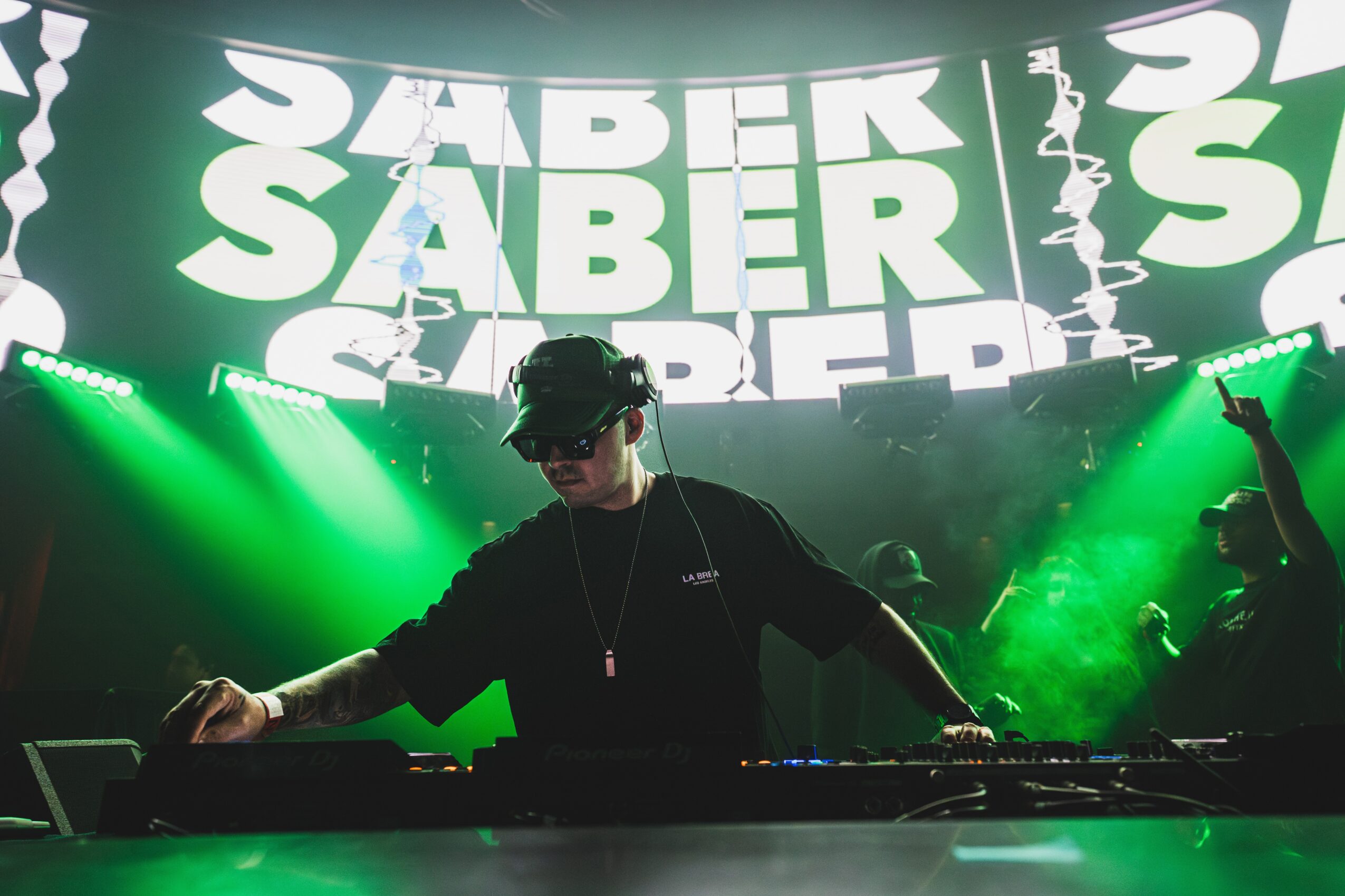 DJ wearing headphones and sunglasses performs on stage with green lighting and a large screen displaying text in the background.