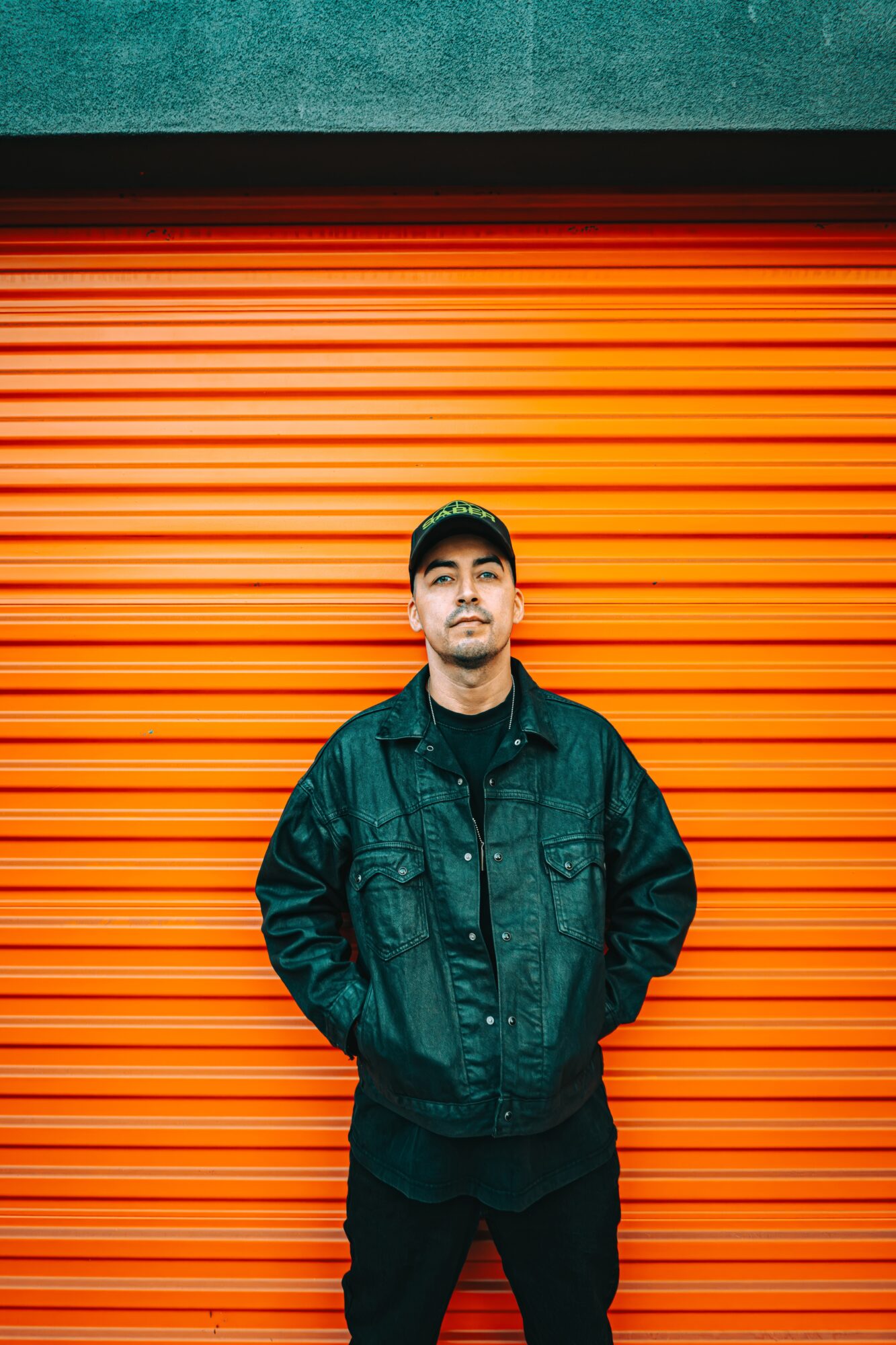 Young man in black jacket and cap stands in front of orange rolling shutter door, hands in pockets.