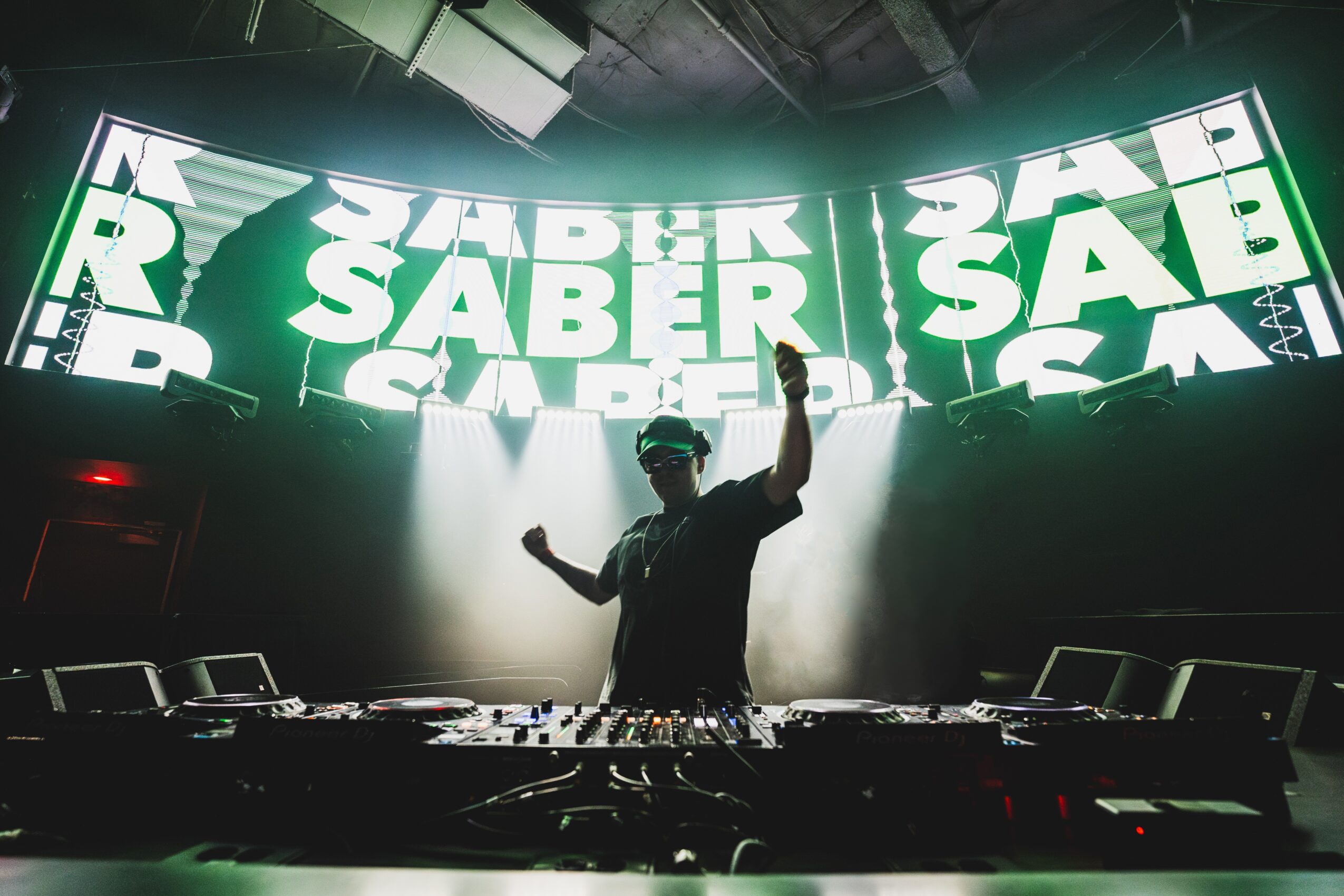 DJ performs on stage with large digital screens displaying text in the background, raising one hand, in a dark setting.