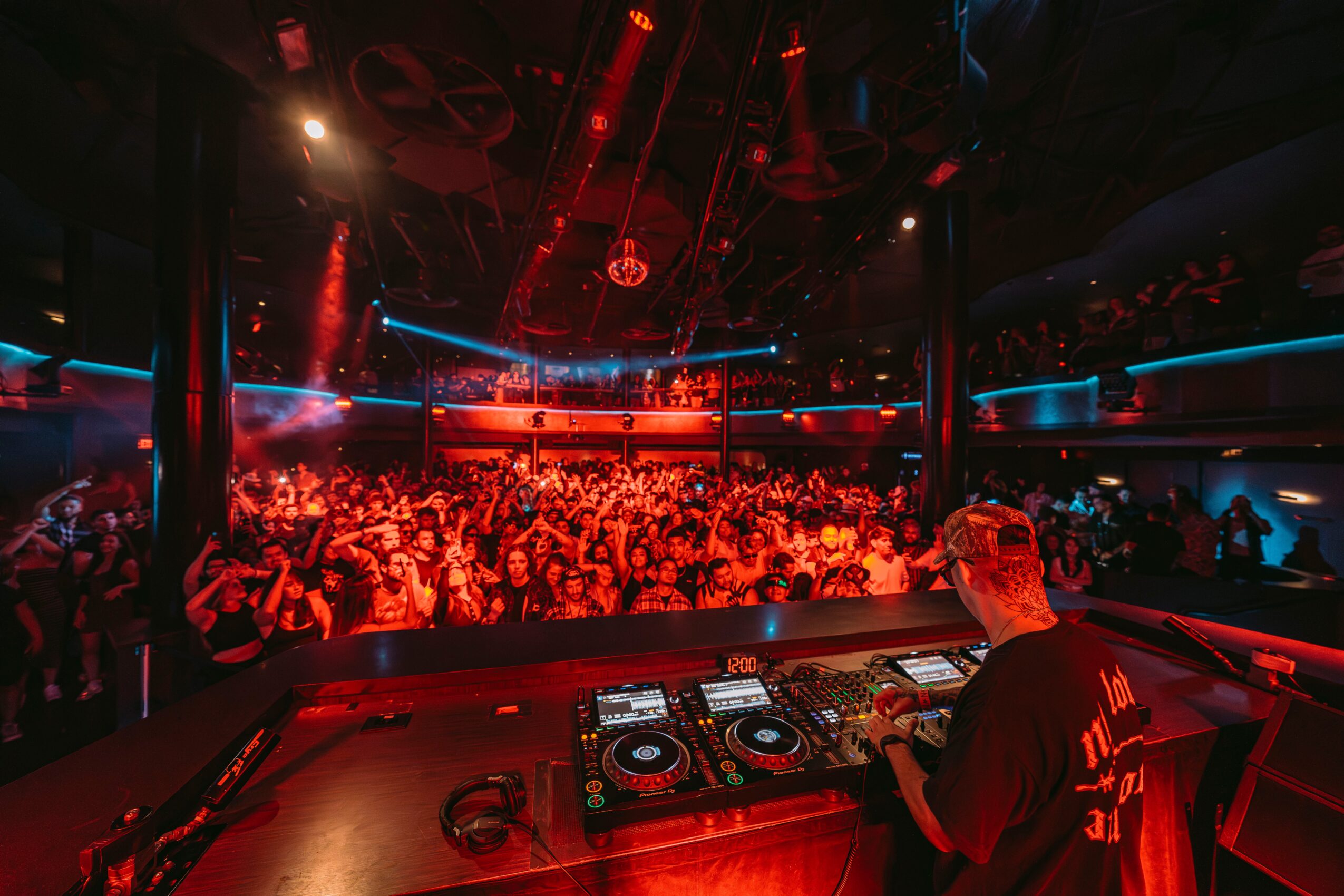 DJ performing at a nightclub with a large crowd, colorful lights, and a dance floor, viewed from behind the DJ booth.